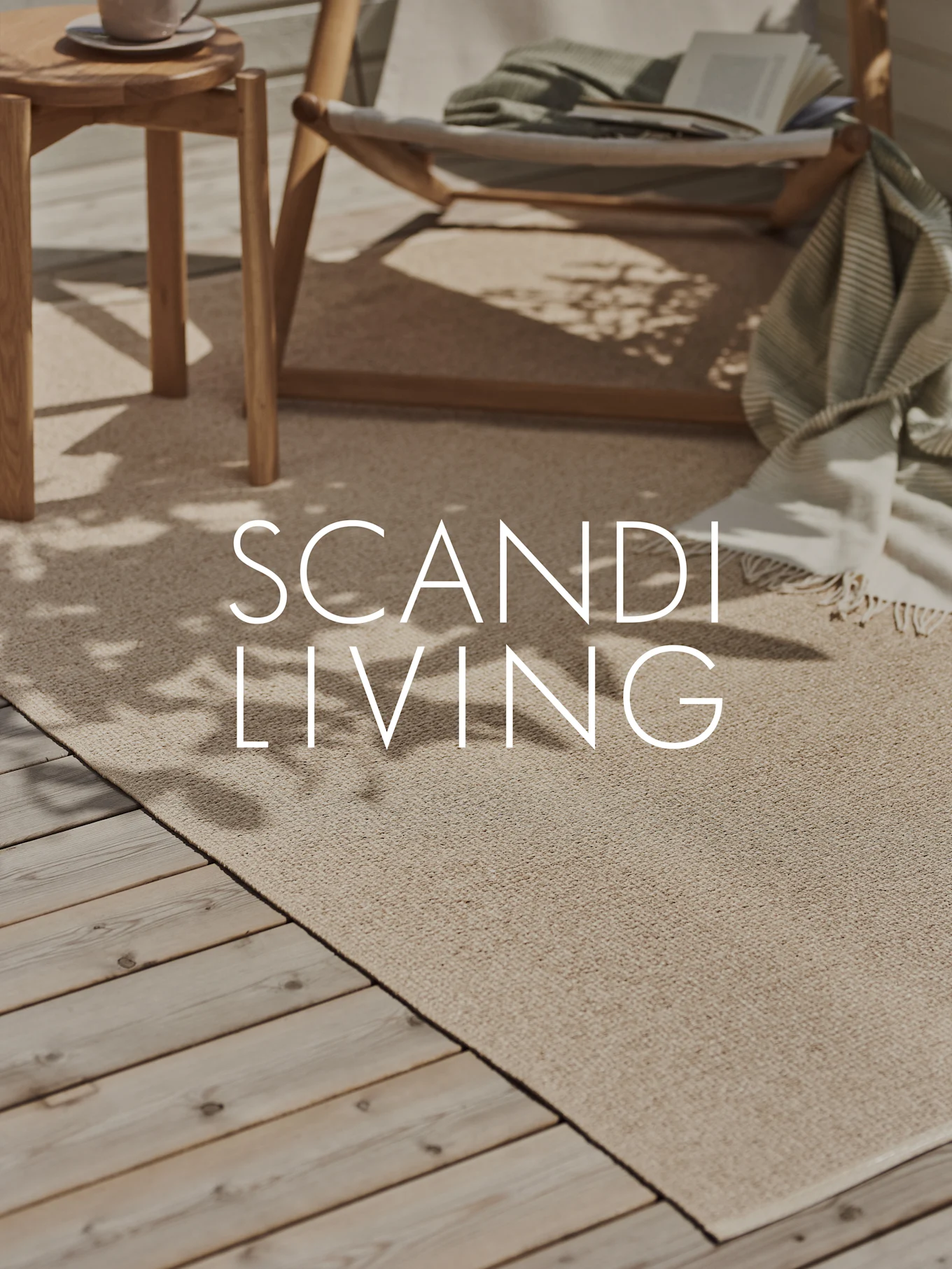 Una alfombra beige claro para exterior en una terraza de madera con una tumbona, una manta, un libro y una mesa auxiliar con una taza. Texto: SCANDI LIVING.