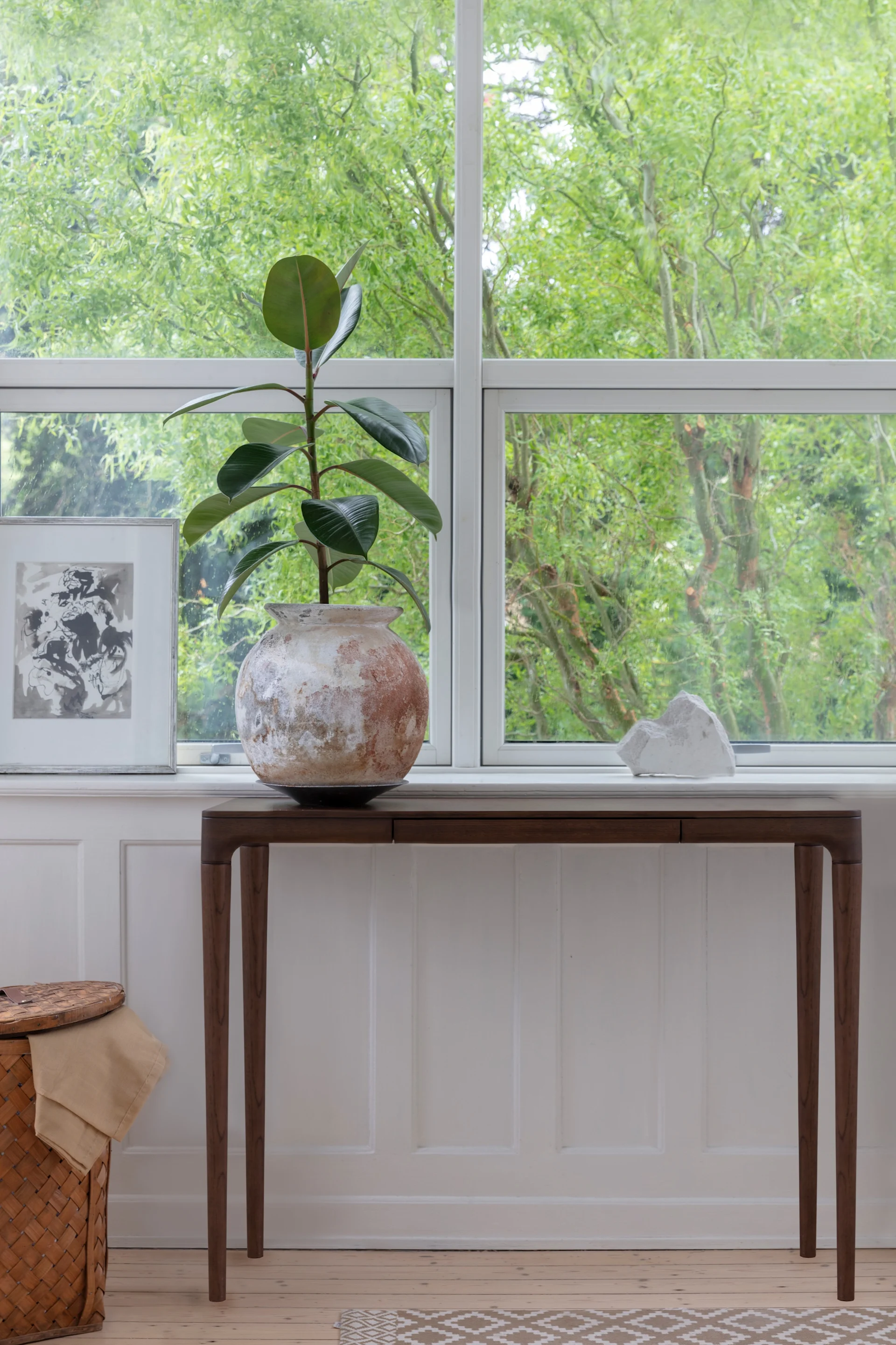 Una planta de caucho en una maceta rústica sobre una mesa consola junto a una ventana, con una obra de arte enmarcada y una escultura blanca. Se ven árboles verdes afuera.