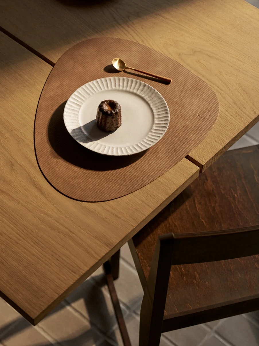 Desde arriba: canelé en plato blanco, cuchara dorada sobre mantel marrón en mesa de madera.