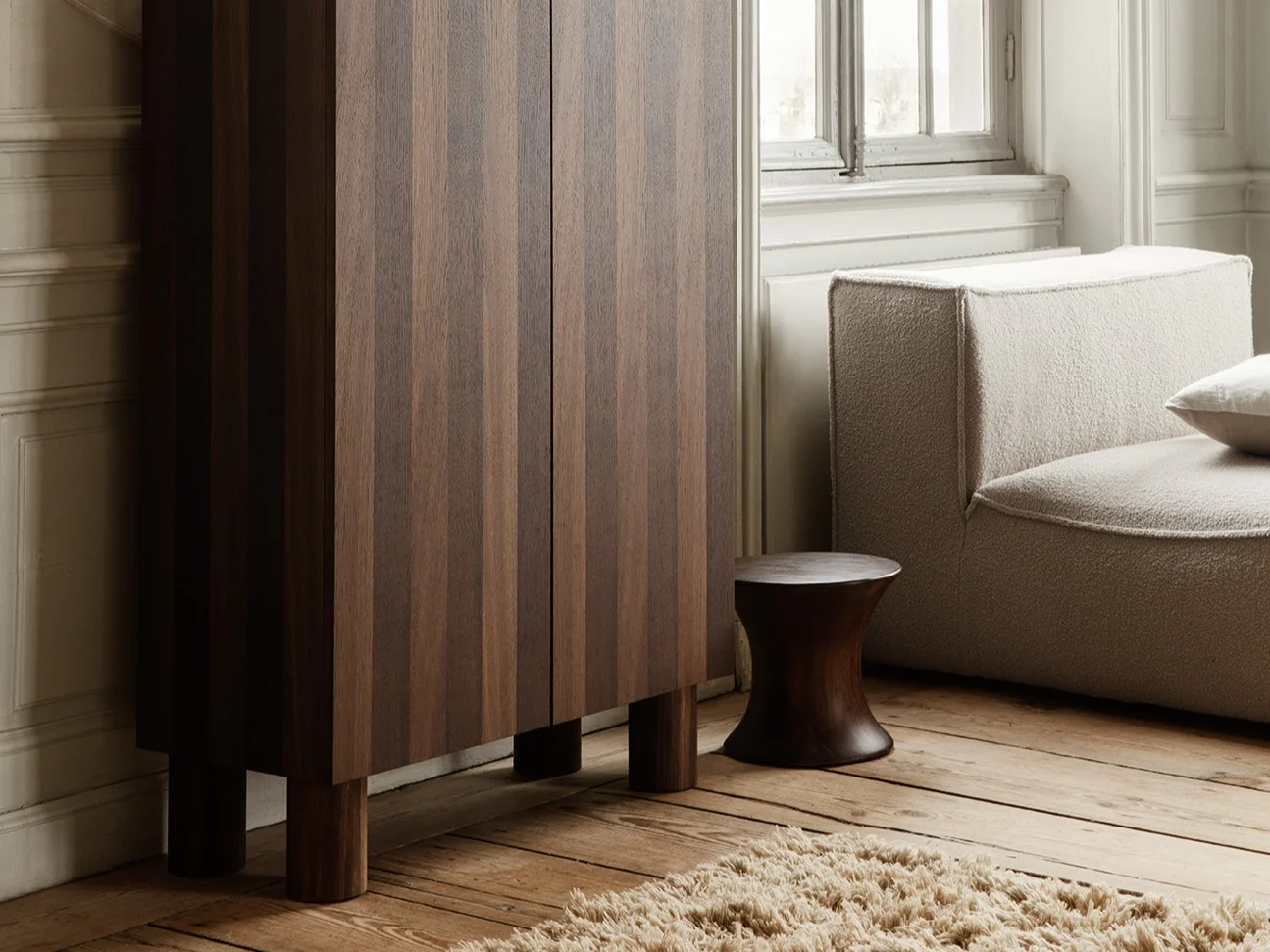 Elegante habitación con armario de madera oscura estriado, escultura de mujer arrodillada, sofá modular claro y gran ventana.
