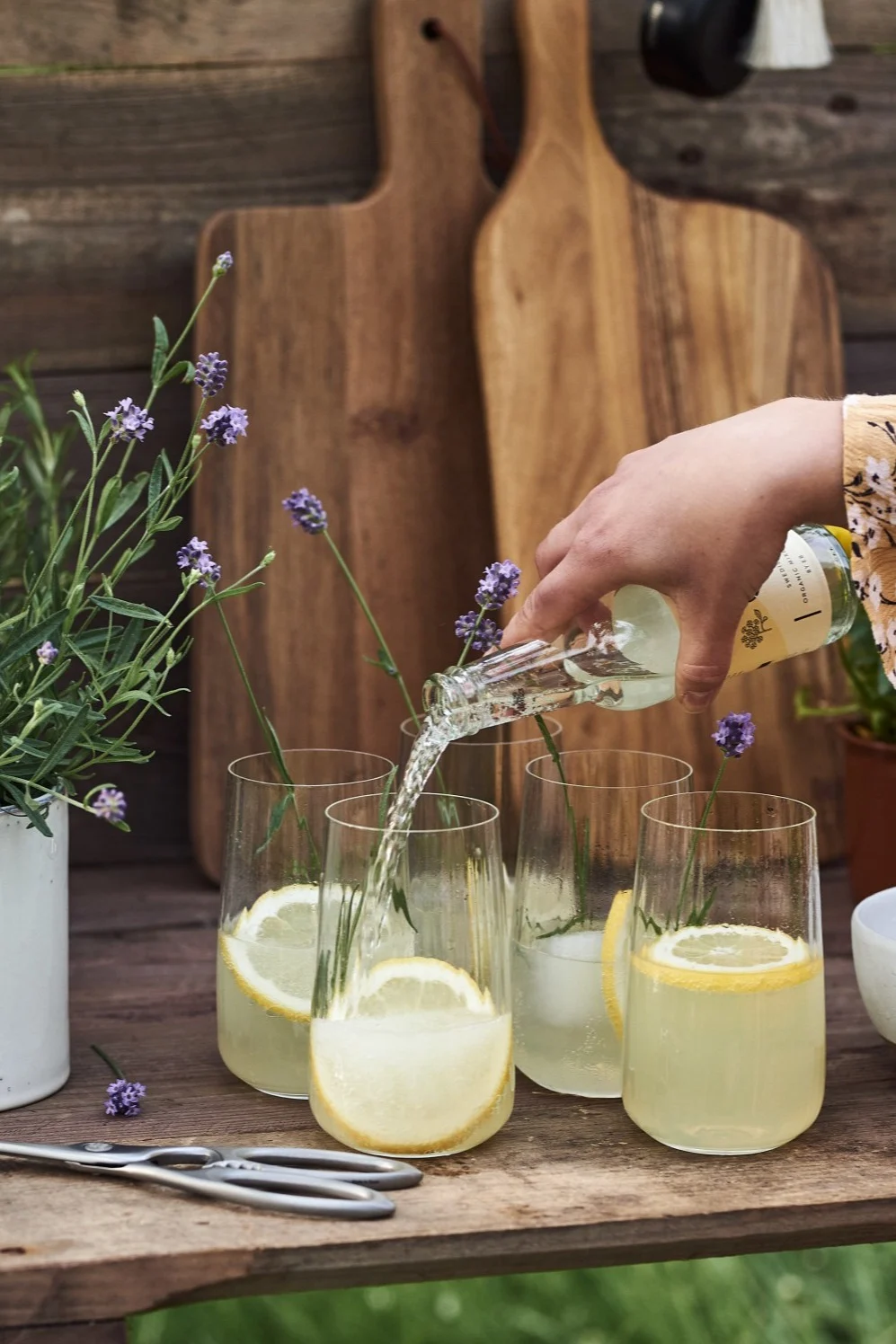 Una persona sirviendo una bebida de una botella pequeña en vasos con rodajas de limón y lavanda sobre una mesa de madera.