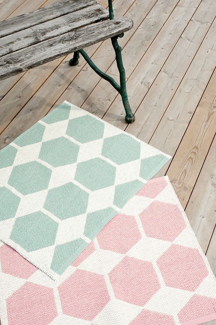 Alfombra de exterior: aquí puedes ver la alfombra de exterior verde y rosa de Brita Sweden colocada delante de un banco en una terraza de madera.