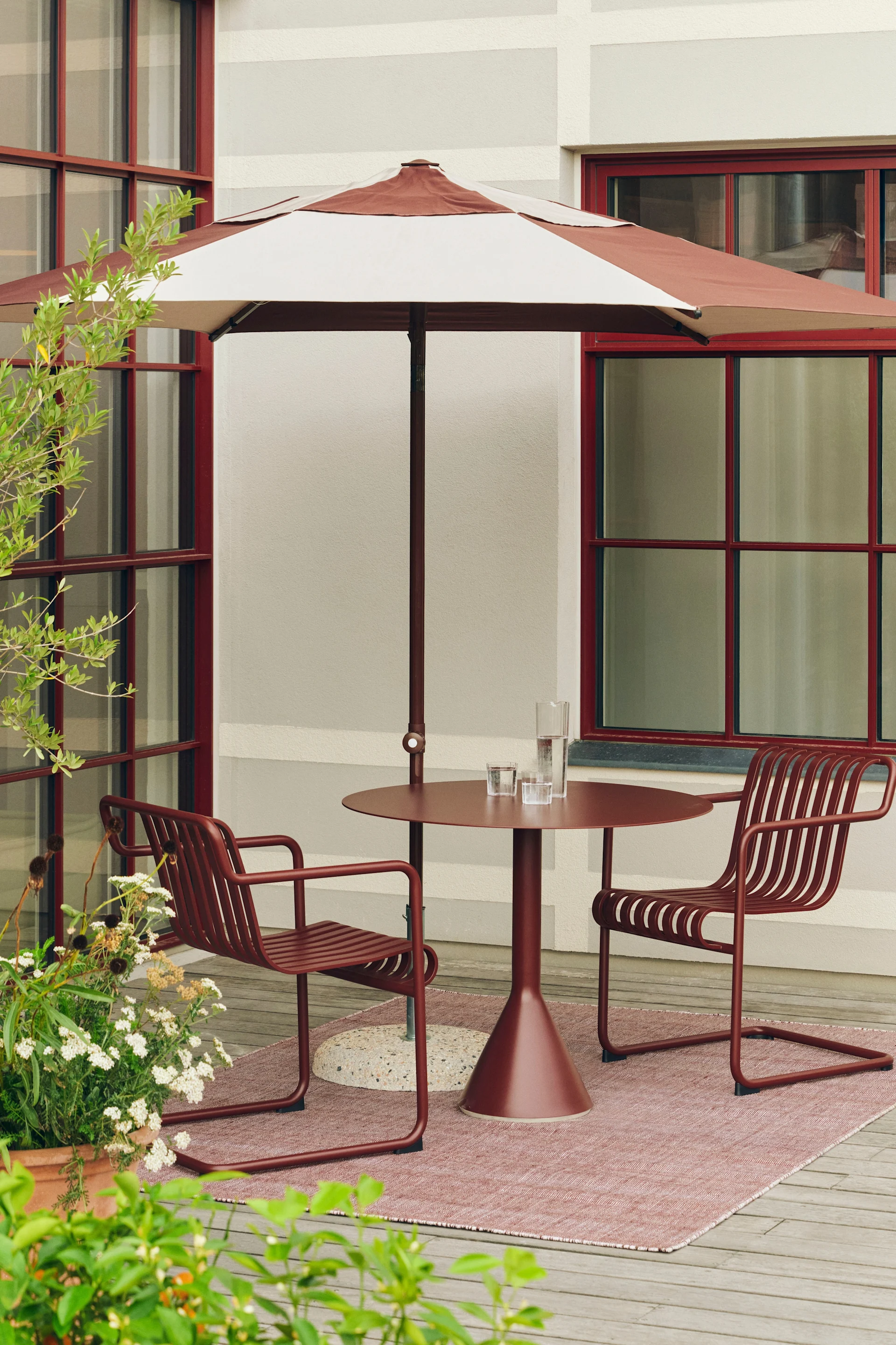 Patio exterior con sombrilla rayada rojo-marrón, mesa redonda a juego y dos sillas con lamas sobre una alfombra rosa.