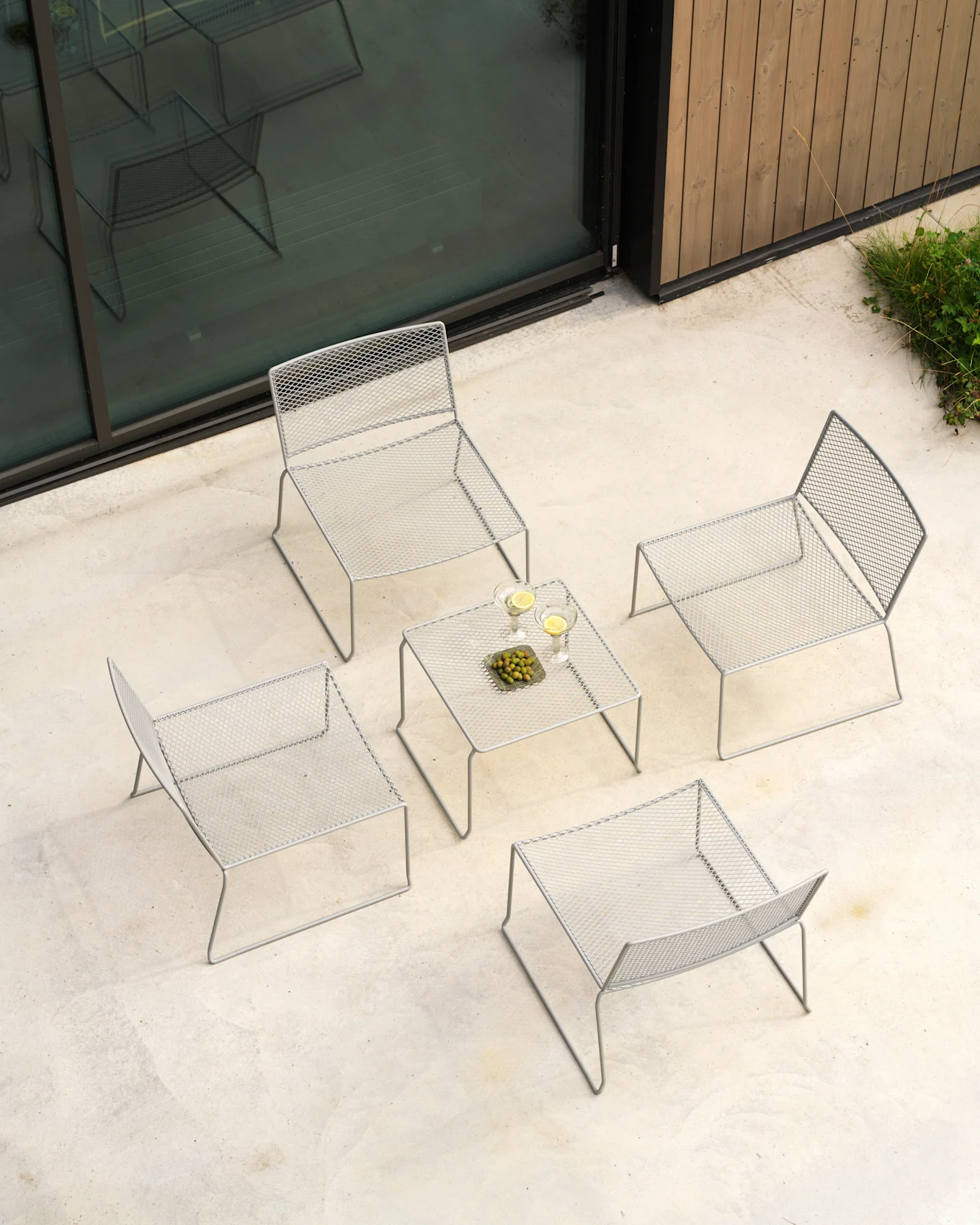 Vista aérea de un set lounge gris claro: cuatro sillas de malla, mesa con bebidas y aceitunas en patio de hormigón.