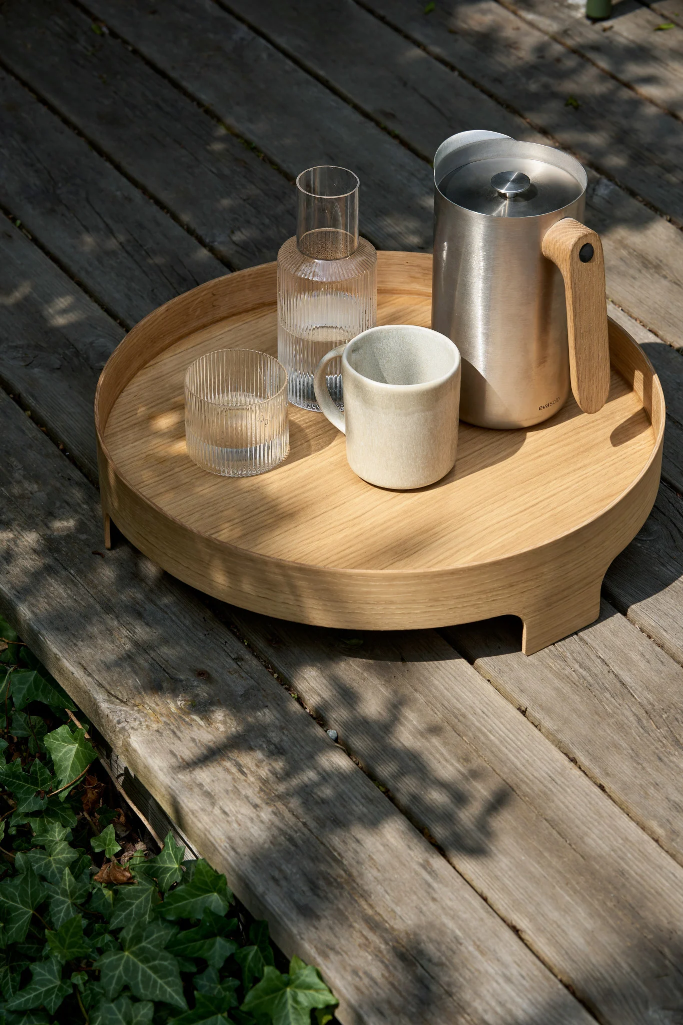 Una bandeja redonda de madera en una terraza con una jarra de acero inoxidable con mango de madera, una garrafa de cristal estriado, un vaso estriado y una taza de cerámica.