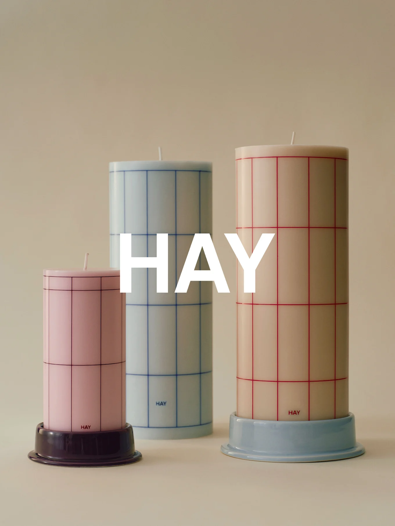 Tres velas cilíndricas con patrón de cuadrícula de diferentes alturas: rosa, azul claro y crema, sobre bases.