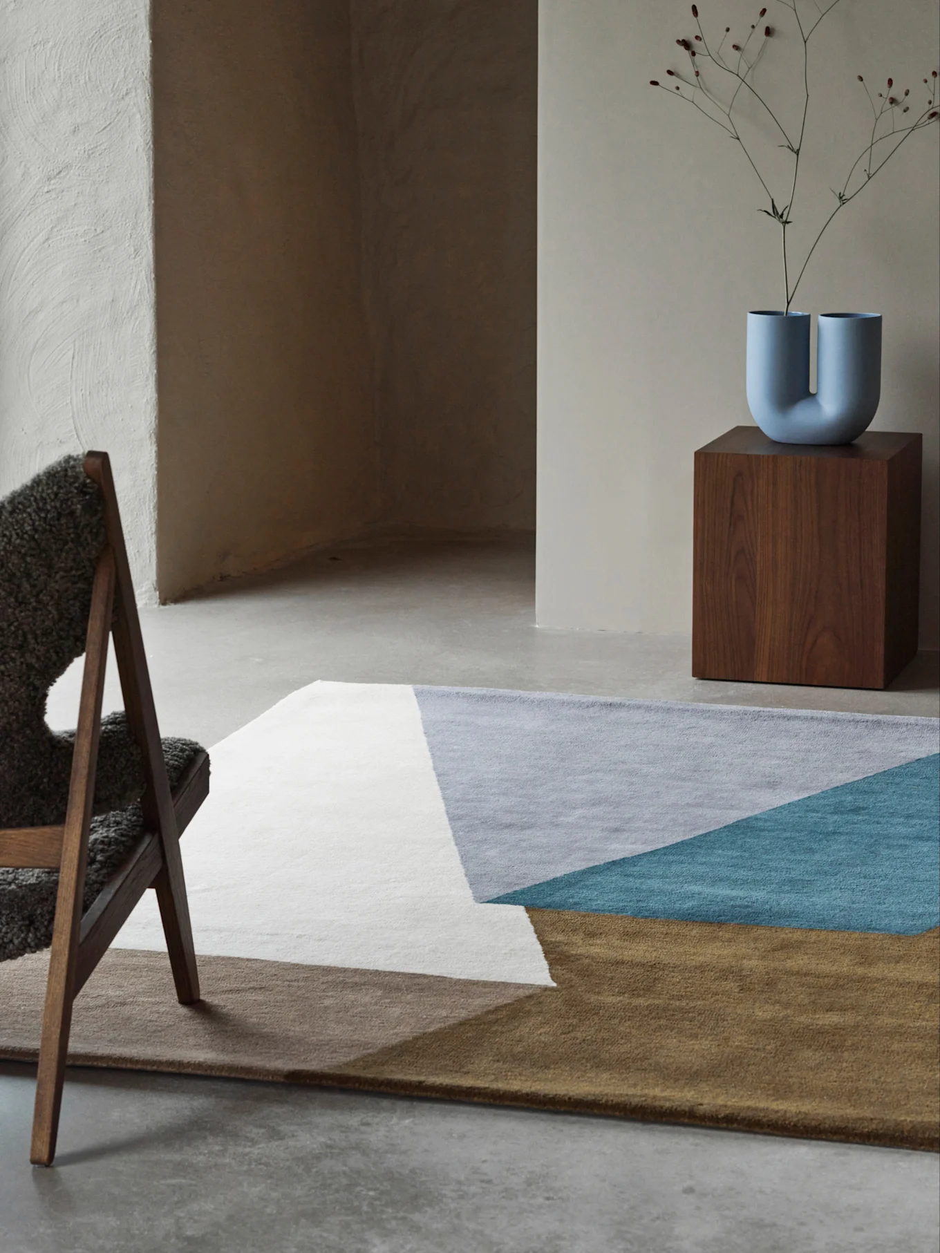 Habitación moderna con alfombra geométrica en blanco, gris, turquesa y marrón, un jarrón azul en forma de U sobre un pedestal de madera y una silla mullida.