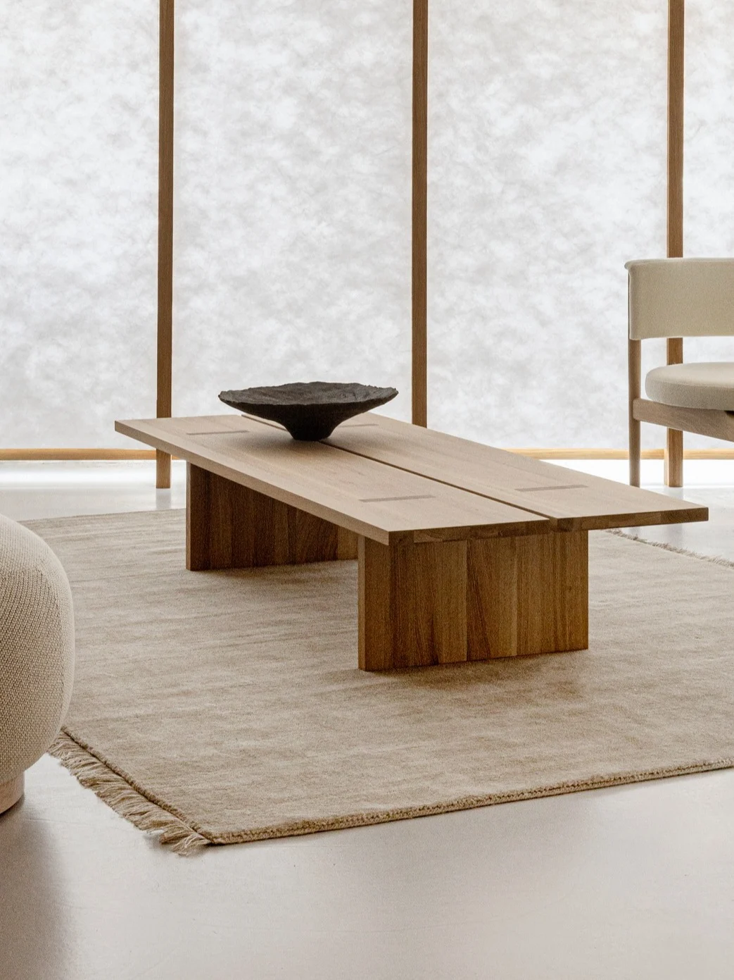 Sala de estar minimalista con mesa de centro de madera, alfombra beige, sillón y silla frente a pantallas de papel.