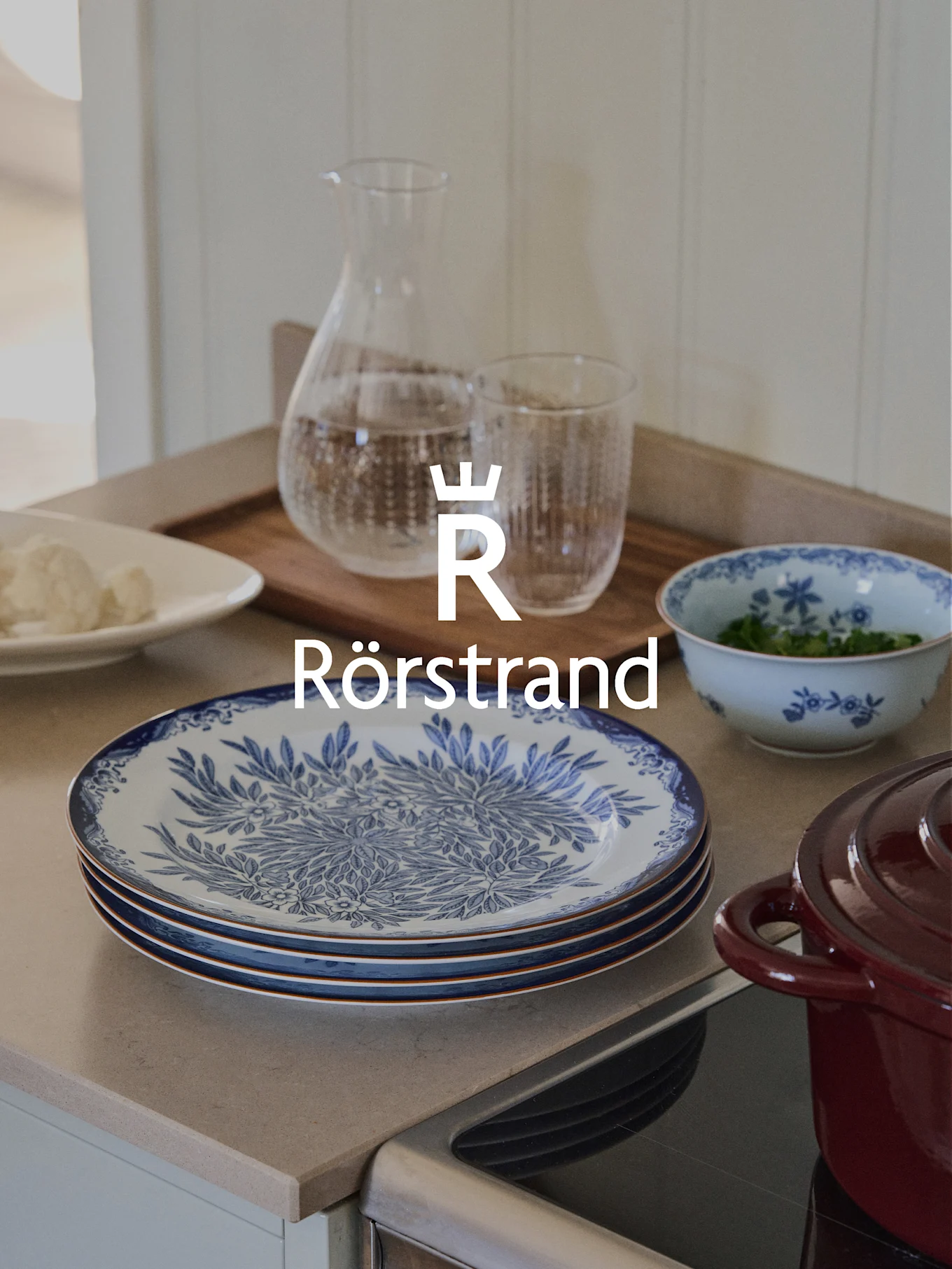 Logo de Rörstrand sobre encimera de cocina con platos azules y blancos, jarra, vaso y olla roja.