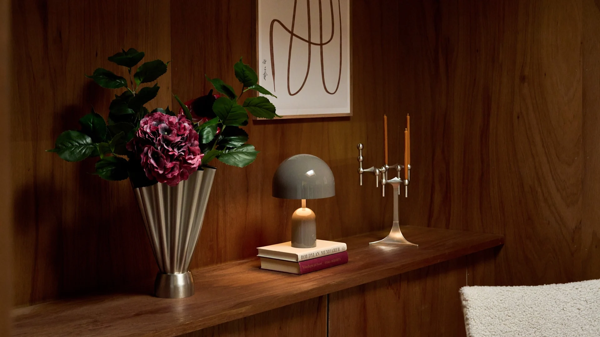Florero con flores rosas oscuras, lámpara gris de hongo sobre libros, candelabro plateado en estante de madera con arte.