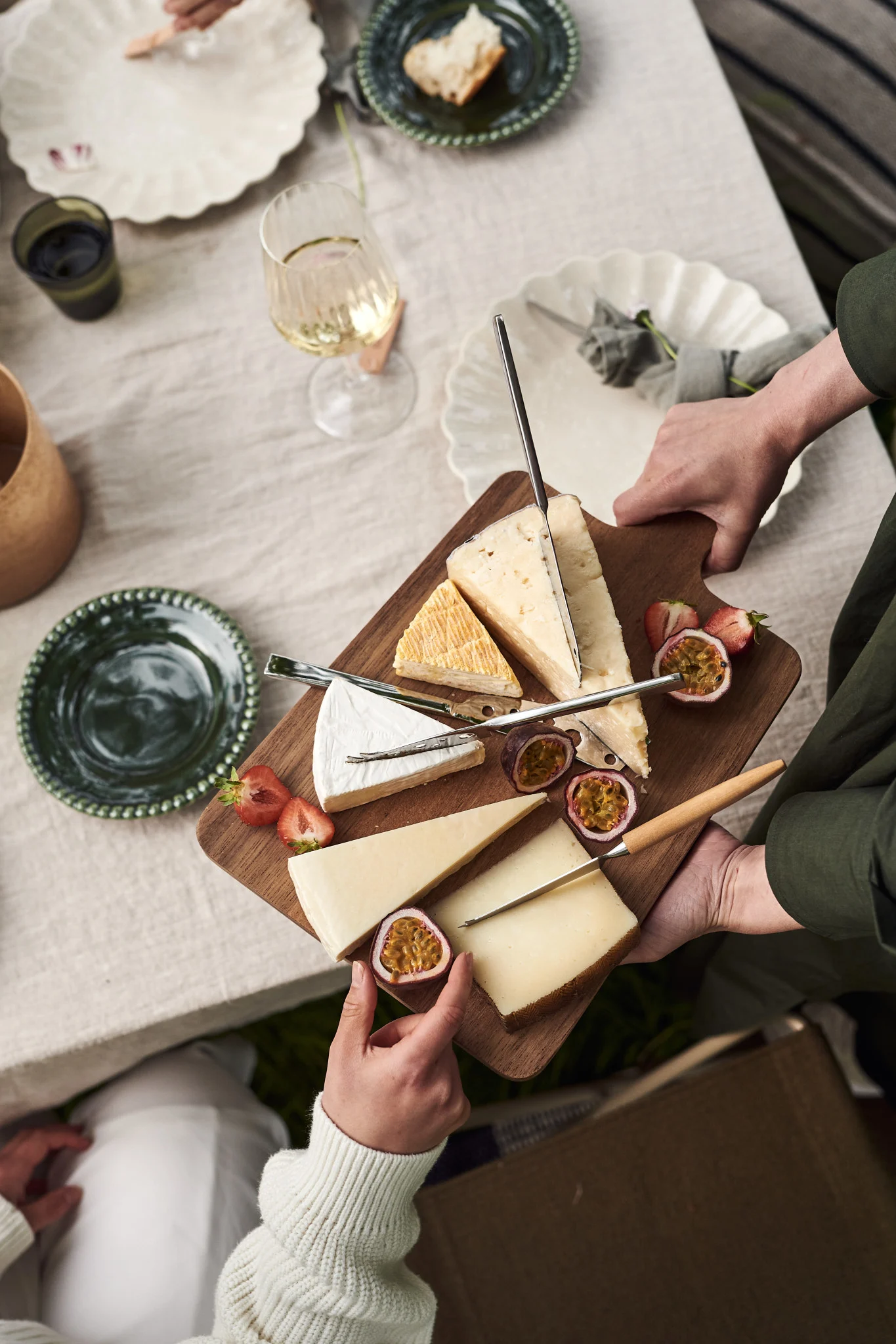 Manos sirven una tabla de quesos de madera con quesos variados, fresas partidas y fruta de la pasión en una mesa.