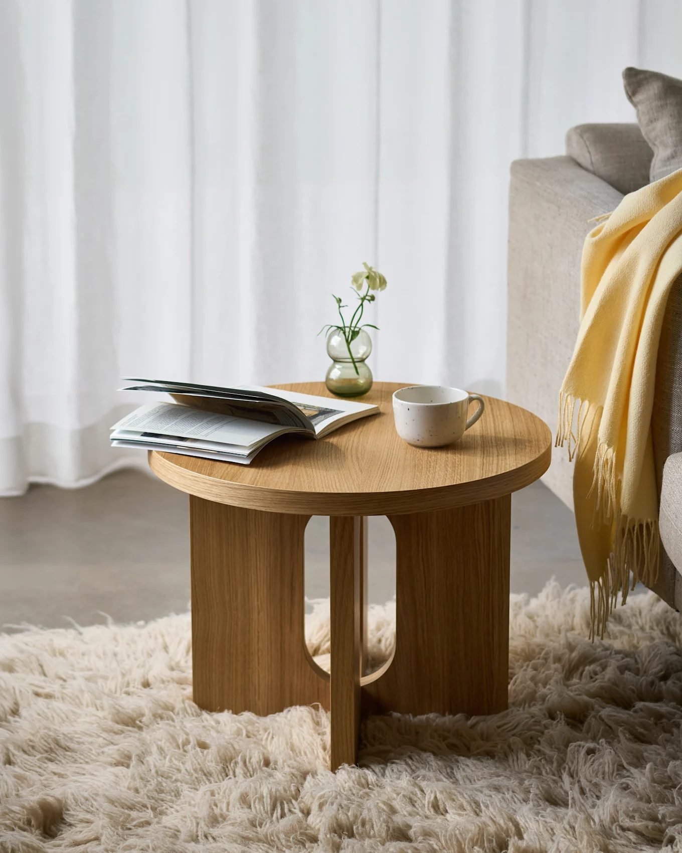 Mesa auxiliar redonda de madera sobre una alfombra mullida, con una revista abierta, una flor blanca en un jarrón y una taza de cerámica, junto a un sofá.