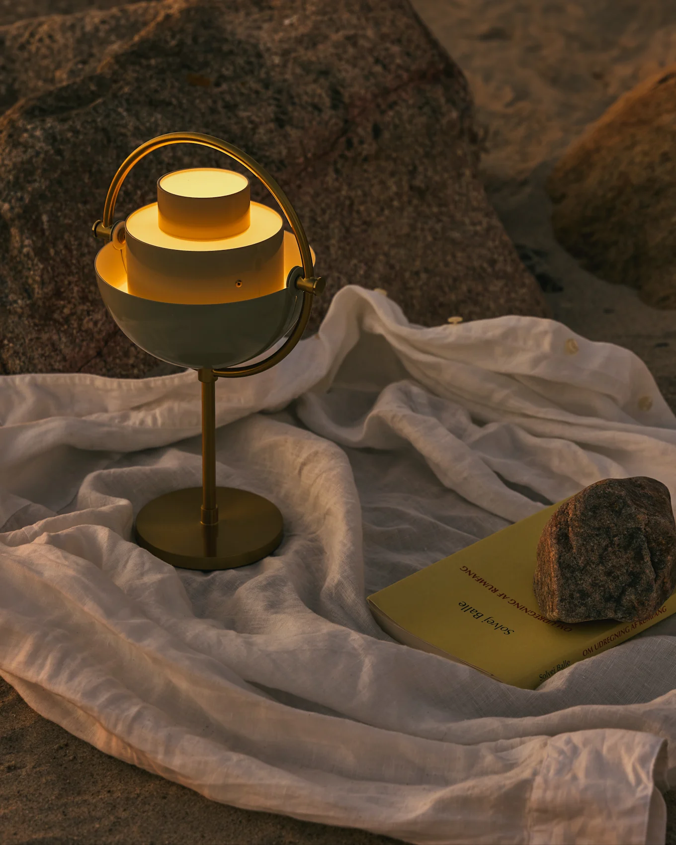 Lámpara de mesa moderna blanca y de latón con luz cálida, un libro amarillo y una roca sobre tela blanca en una superficie rocosa.