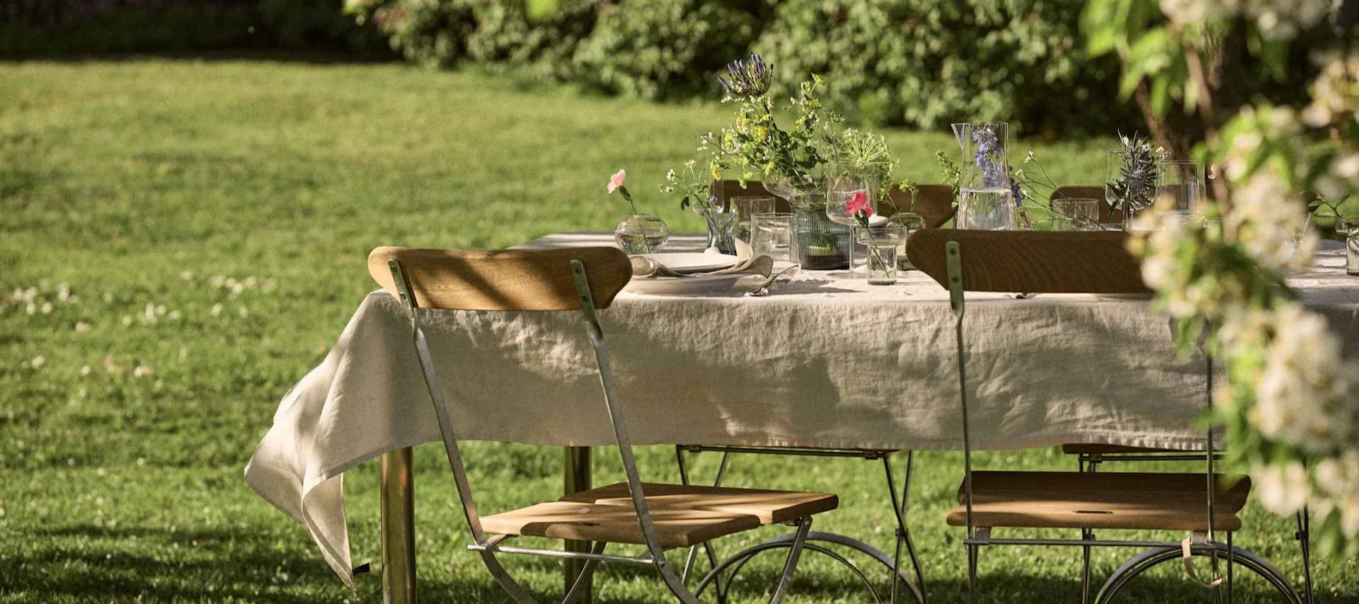 Una mesa de comedor al aire libre puesta con un mantel de lino, sillas de madera, vasos, platos y jarrones de flores silvestres en un césped verde.