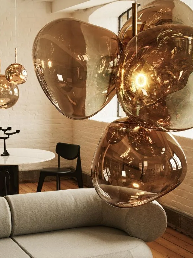 Una sala de estar moderna con un sofá curvo gris, un puf a juego con revistas y lámparas colgantes de cristal ámbar.