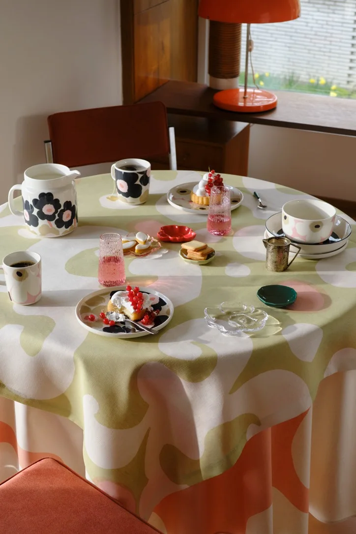 Una mesa redonda con un mantel de flores en tonos verdes, rosas y blancos, puesta con tazas, platos y una jarra con motivos en negro y rosa, pasteles decorados con grosellas rojas y vasos rosas.