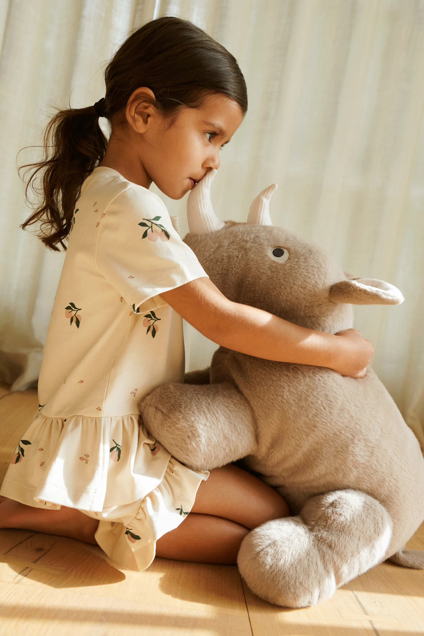Niña con vestido estampado arrodillada y abrazando un gran juguete de peluche con cuernos.