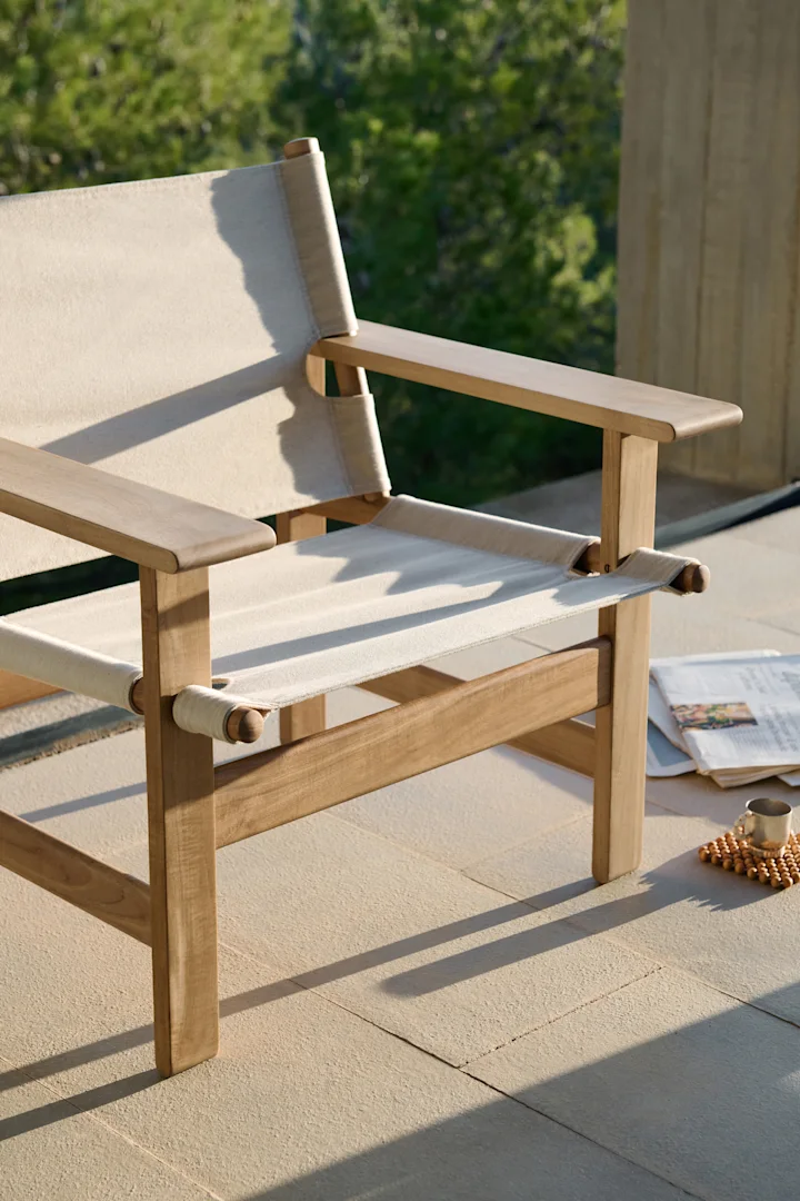 Silla de jardín de madera clara con lona crema en un patio, con un periódico y una taza al lado.