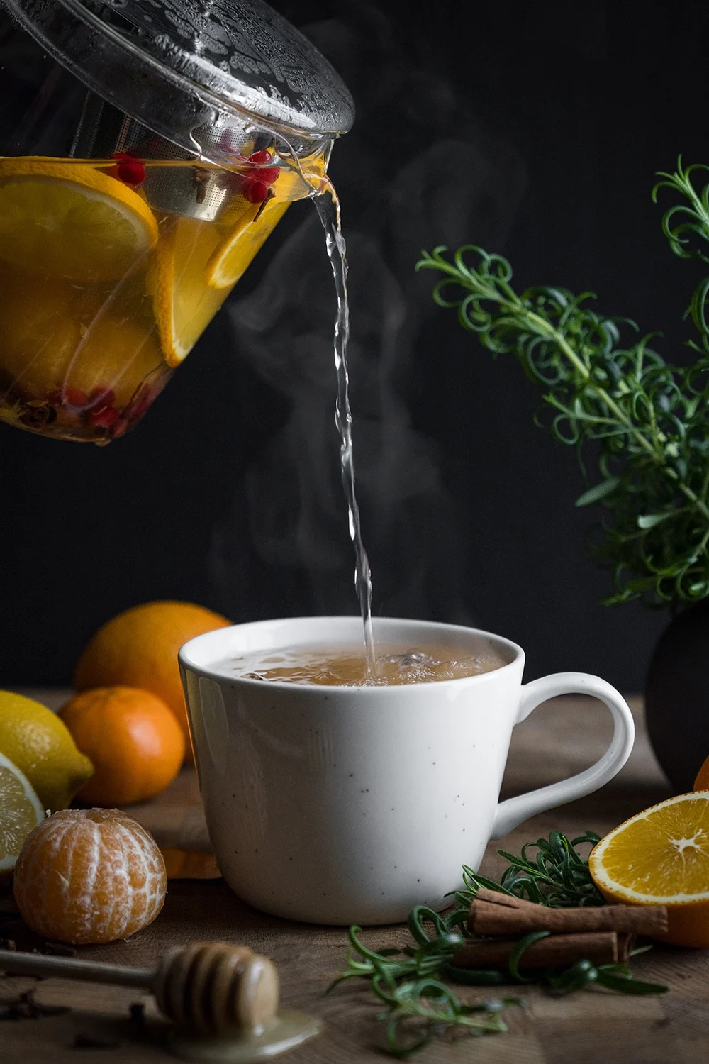 Té caliente con naranjas y bayas vertiéndose de una tetera de cristal en una taza blanca, rodeado de cítricos, romero y canela.