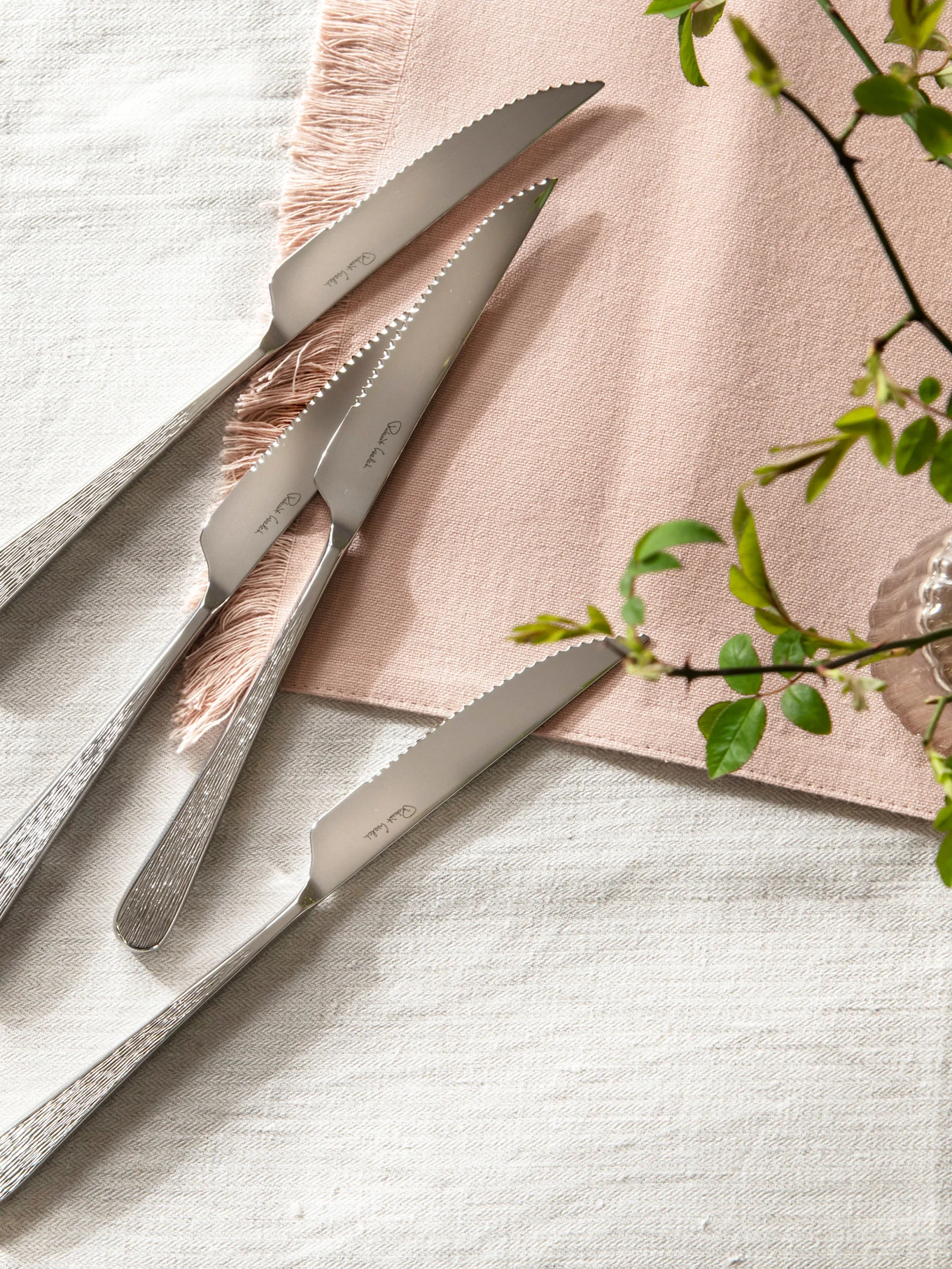 Cuatro cuchillos de carne de acero inoxidable con hojas dentadas y mangos texturizados sobre un mantel de lino con una servilleta rosa.