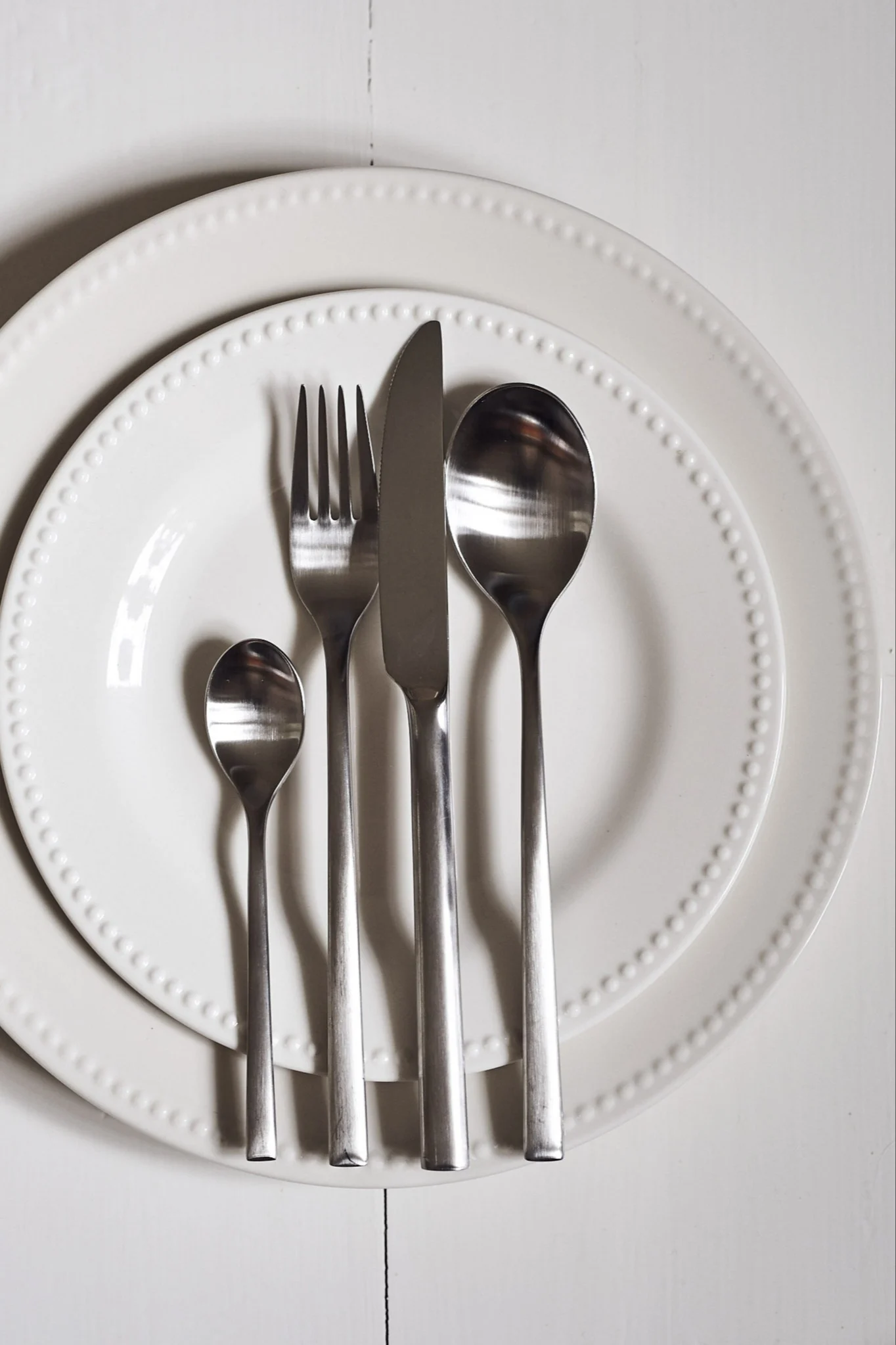 Mesa puesta con dos platos blancos y cuatro piezas de cubertería plateada dispuestas verticalmente.