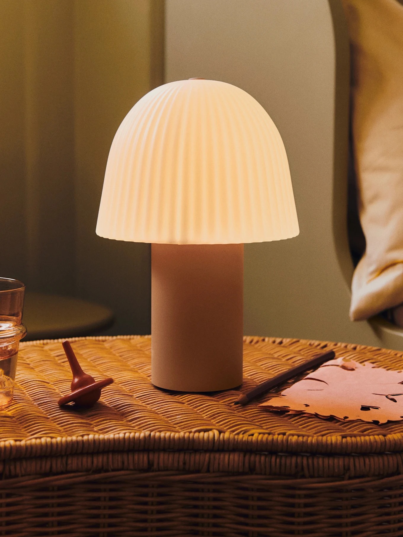 Una lámpara de seta brillante con pantalla plisada sobre una mesita de noche de mimbre con un vaso de agua, una peonza y papel.