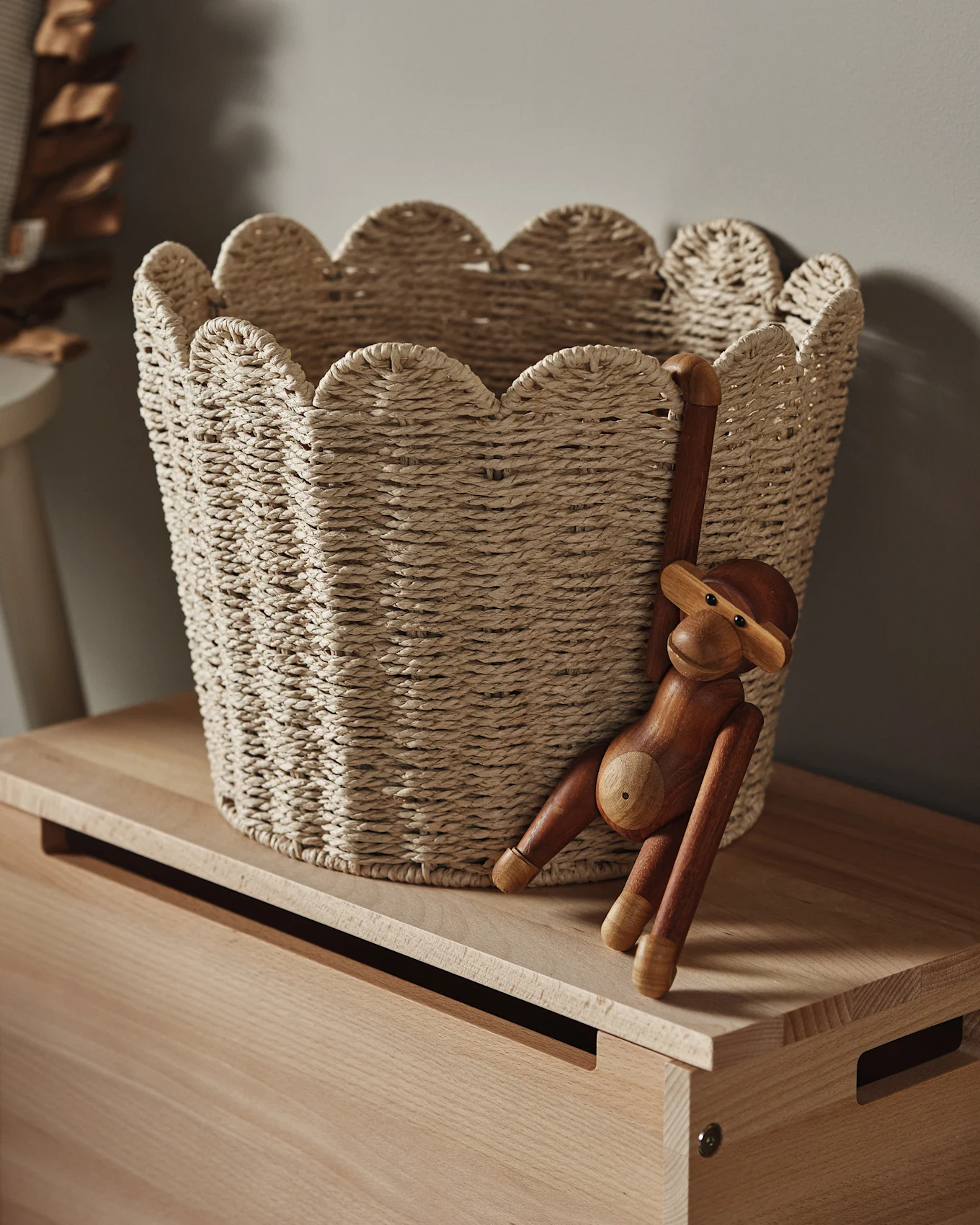 Una cesta de mimbre claro con borde festoneado sobre una superficie de madera, con una figura de mono de madera colgando de su lateral.