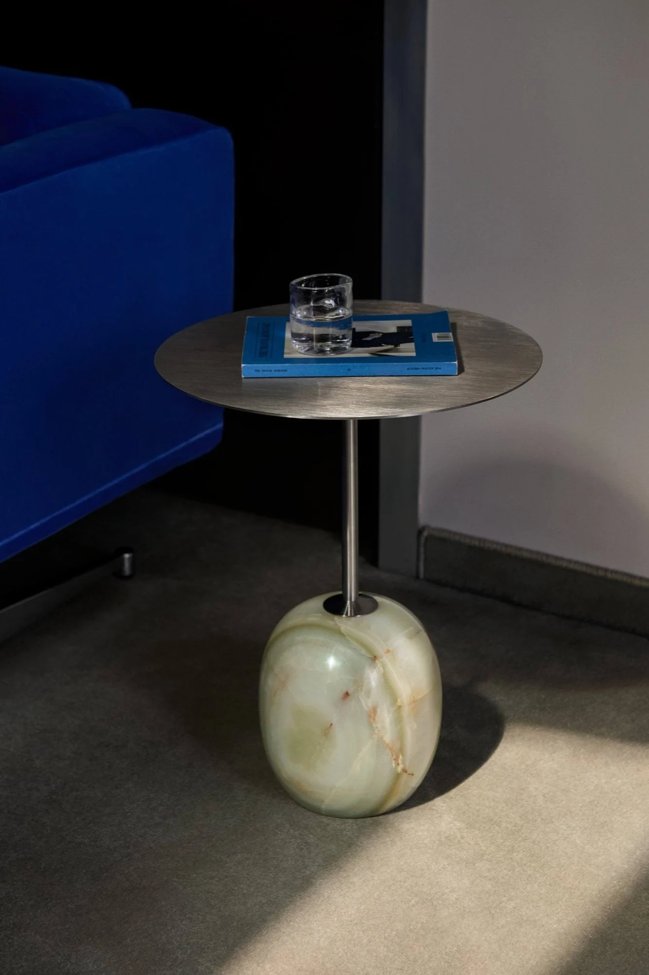 Mesa auxiliar con tapa de metal cepillado, base de ónix verde redondeada, vaso de agua, libro azul, junto a un sofá azul.