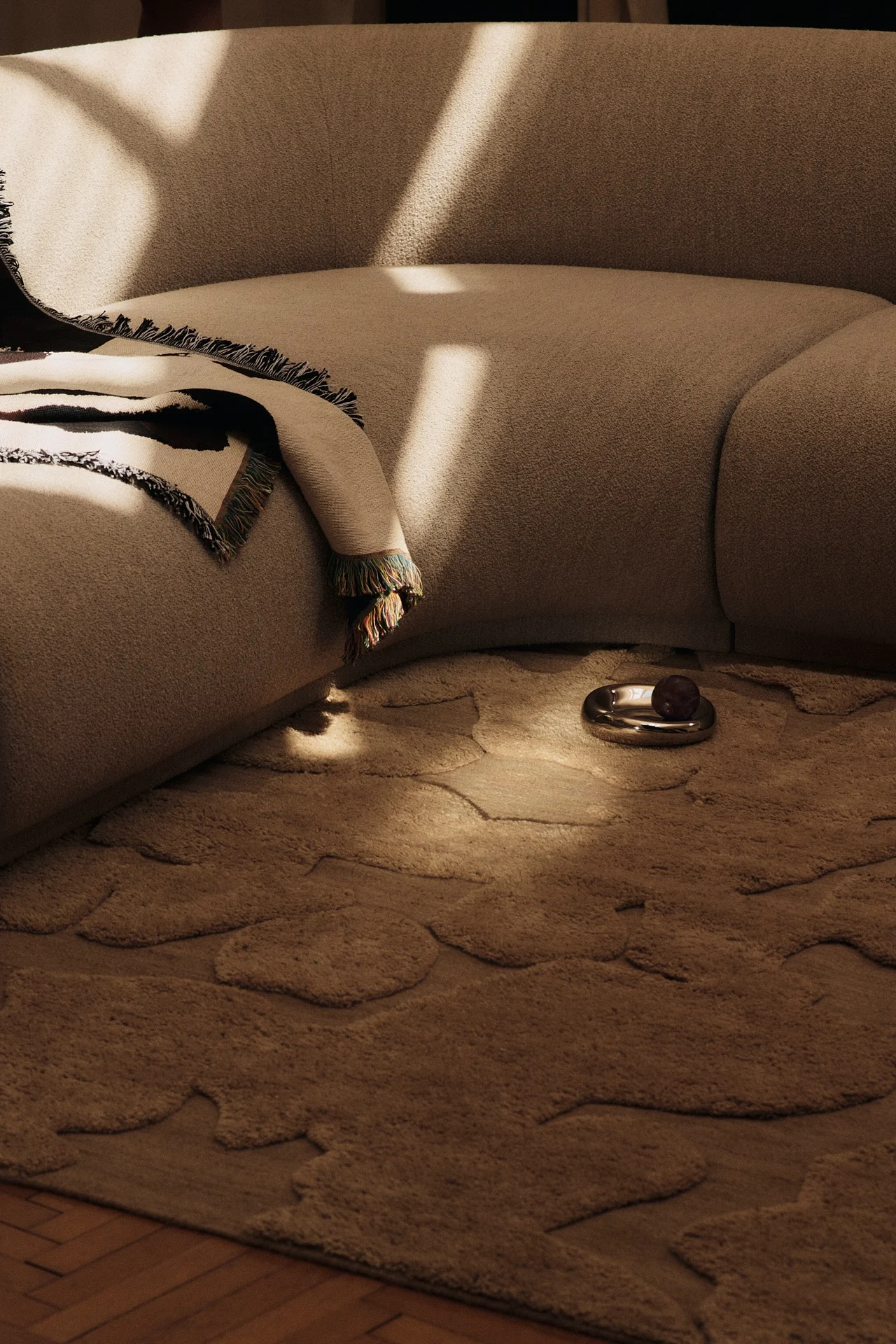 Sofá curvo beige con manta estampada, alfombra beige de forma orgánica y un plato plateado con fruta oscura en una habitación soleada.