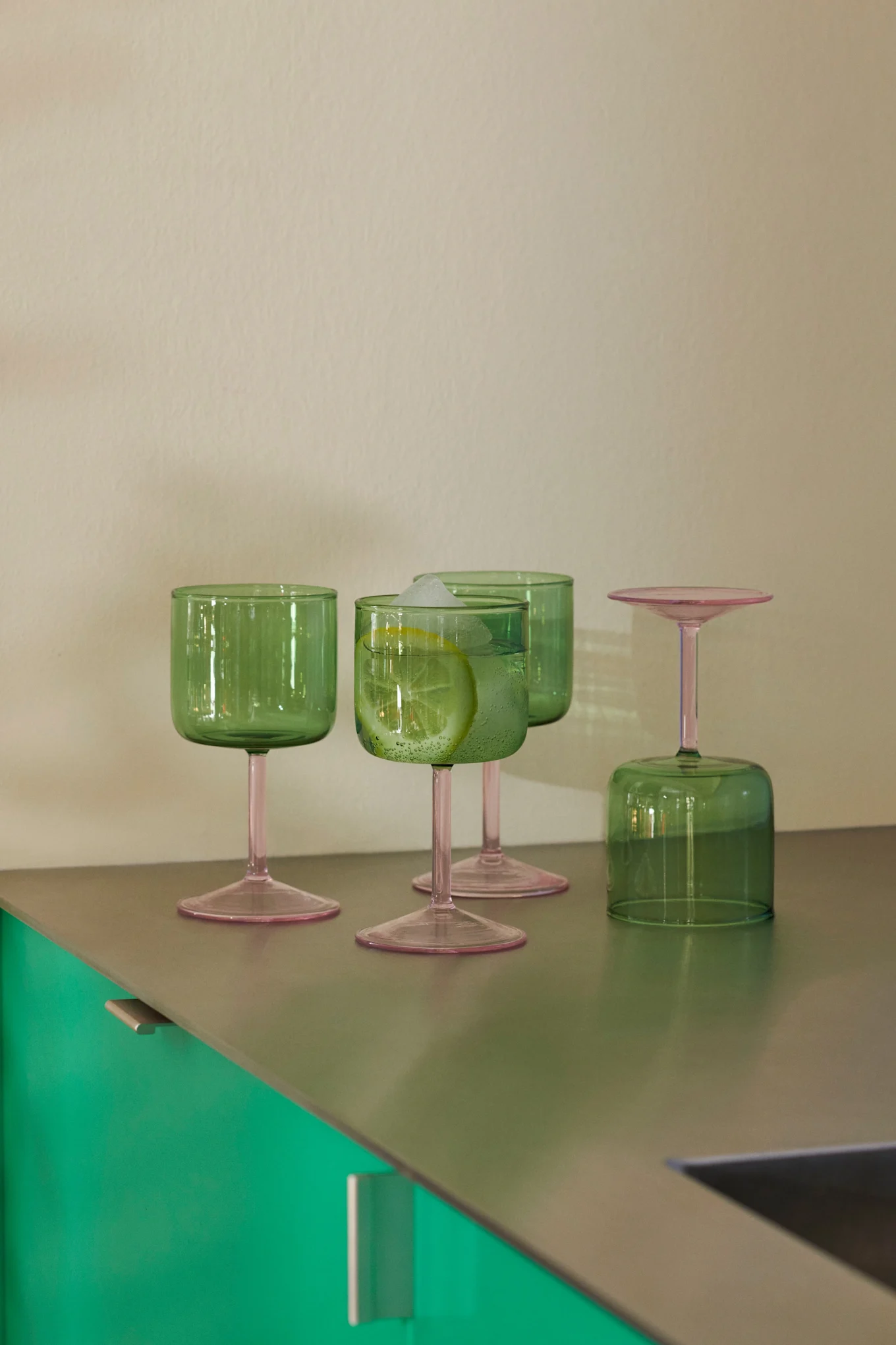 Cuatro copas de cristal verde con tallos rosas sobre una encimera metálica. Una copa contiene una bebida con limón y hielo; otra está del revés.