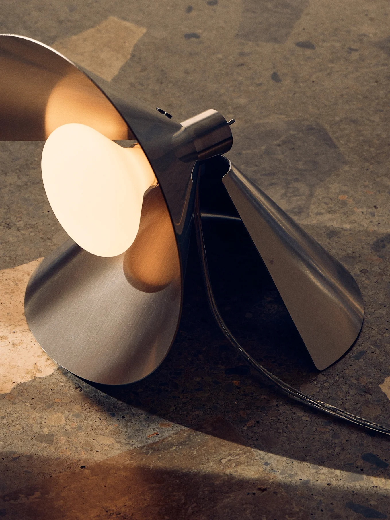 Lámpara de mesa moderna de metal cepillado con diseño escultural plegado y bombilla cálida expuesta sobre superficie texturizada.
