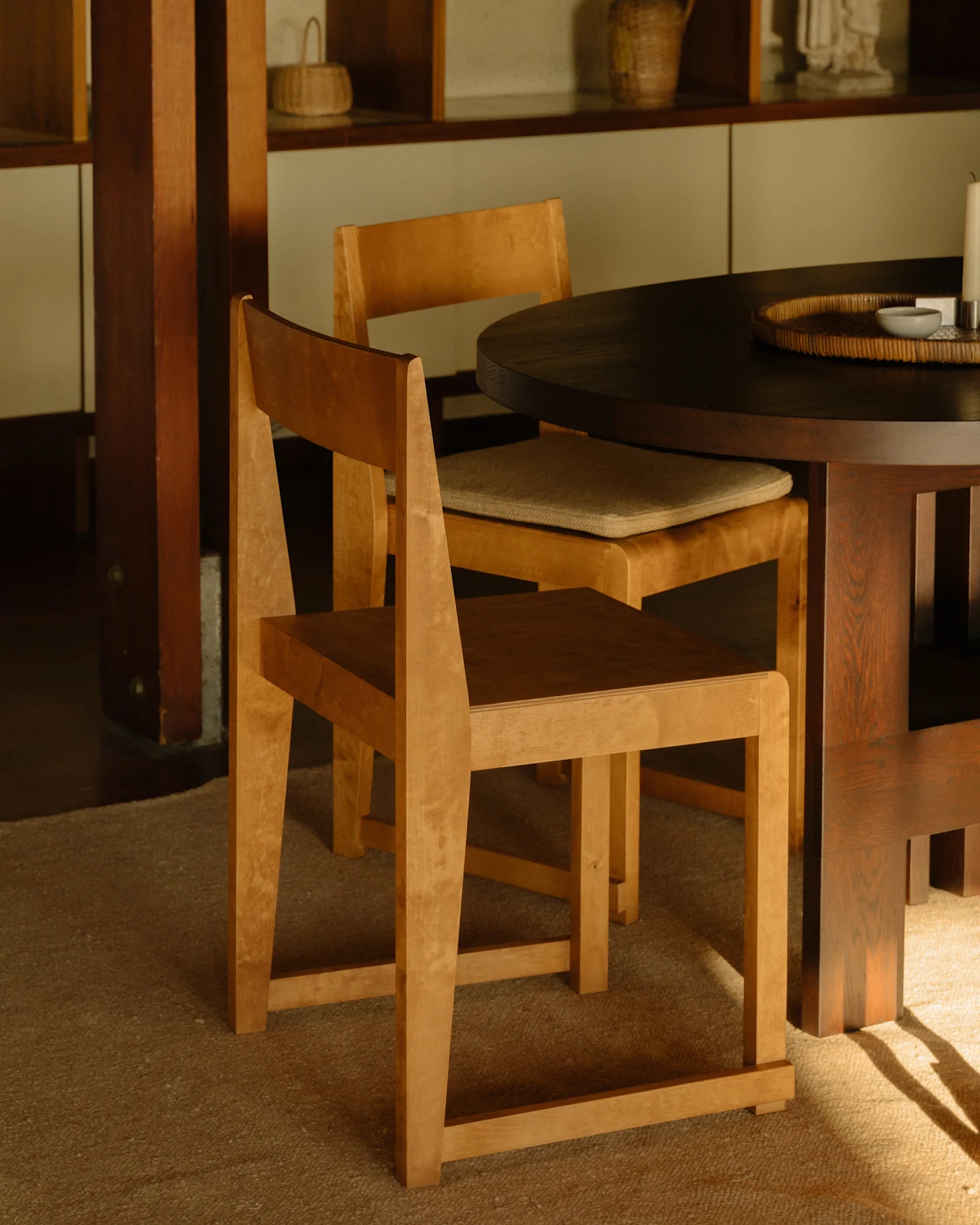 Dos sillas de madera clara en una mesa de comedor redonda oscura, con estanterías de madera al fondo.