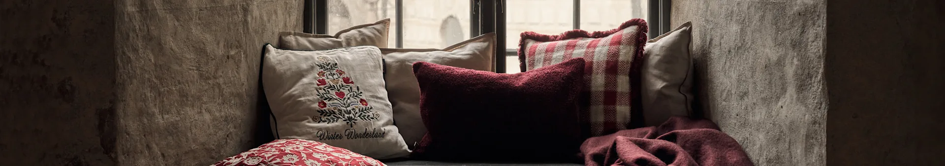 Acogedor asiento junto a la ventana con cojines: uno bordado con un árbol de Navidad, uno rojo texturizado, uno a cuadros rojo y una manta roja.