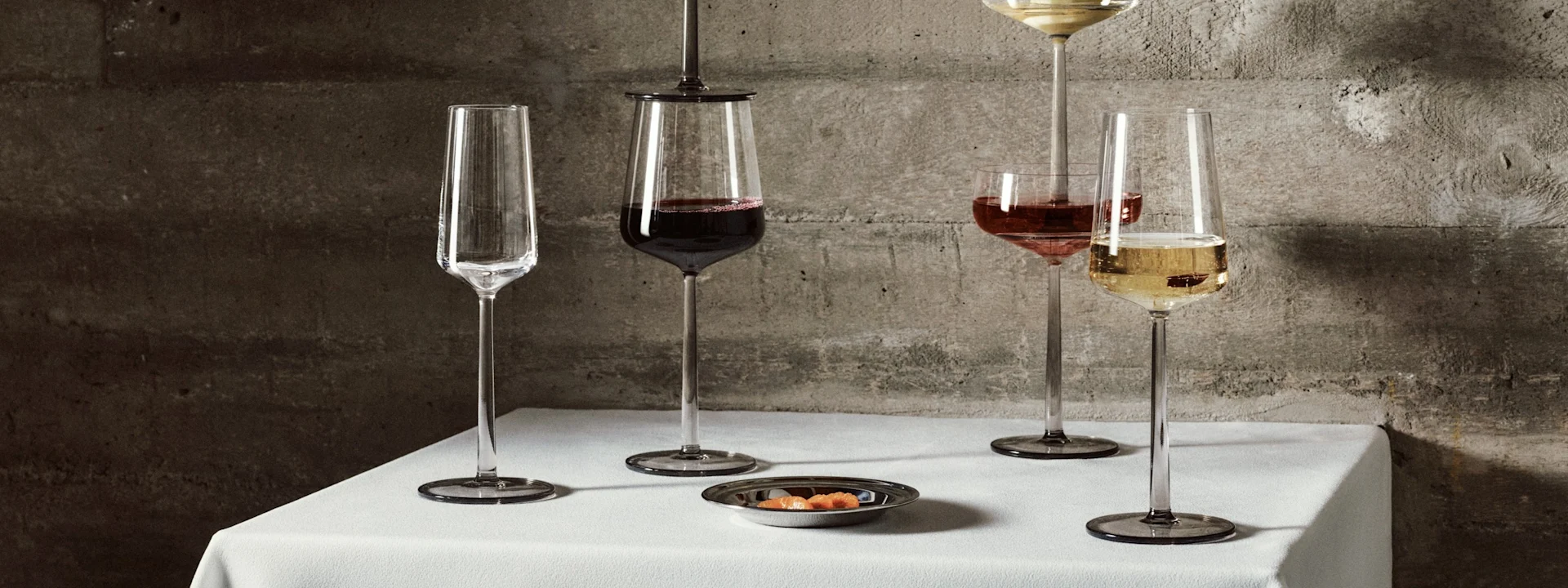 Cinco elegantes copas de tallo largo con vino y champán, una con una pera, sobre un mantel blanco frente a una pared de hormigón.