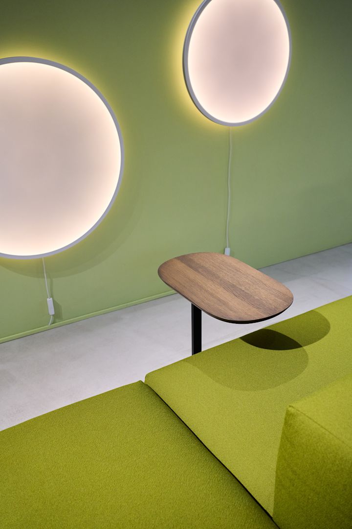 Una habitación con una pared verde de la que cuelgan dos apliques redondos Calma, delante de la cual hay un sofá verde con una mesa auxiliar Relate.