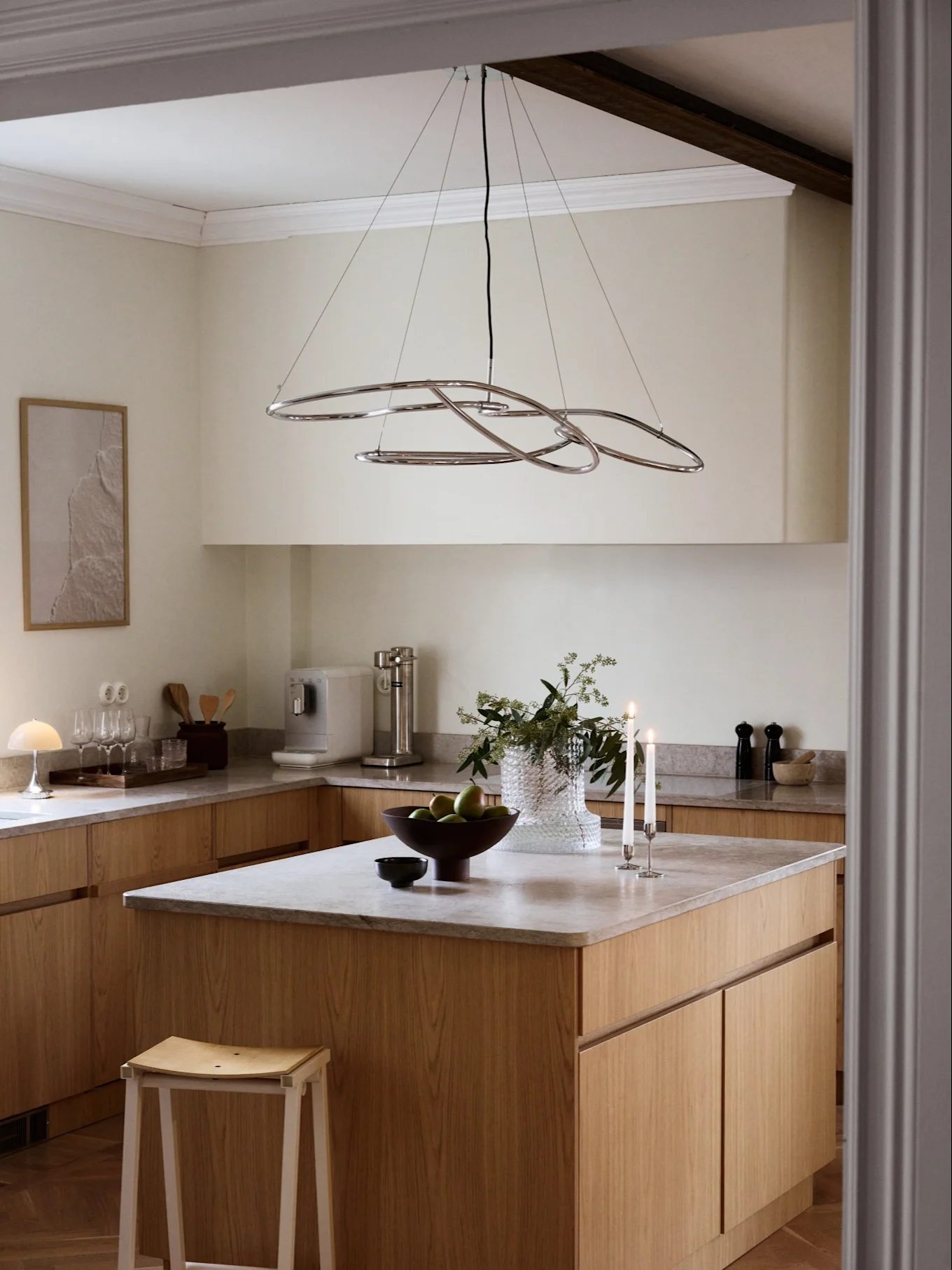 Una cocina moderna con gabinetes de madera clara, encimeras grises y una lámpara de araña abstracta cromada sobre una isla con fruta y velas.
