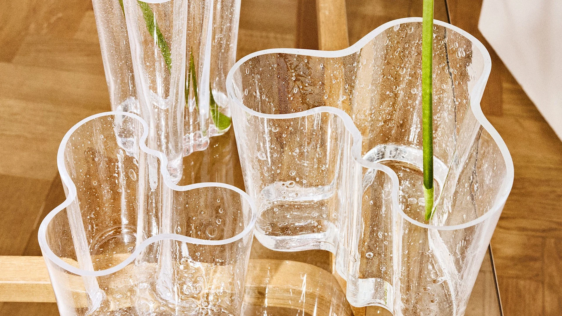 Varias vasijas de vidrio transparente con formas orgánicas onduladas y burbujas de aire incrustadas, con tallos verdes, sobre una superficie de madera.