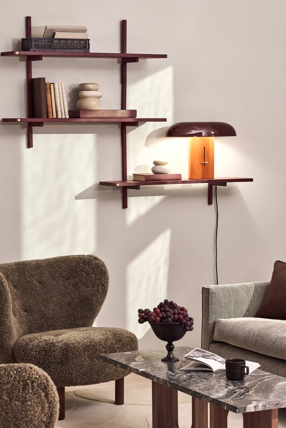 Una sala de estar acogedora con estantes de pared marrones rojizos, una lámpara con pantalla abovedada y base naranja, un sillón marrón texturizado y una mesa de centro de mármol con uvas y un libro.