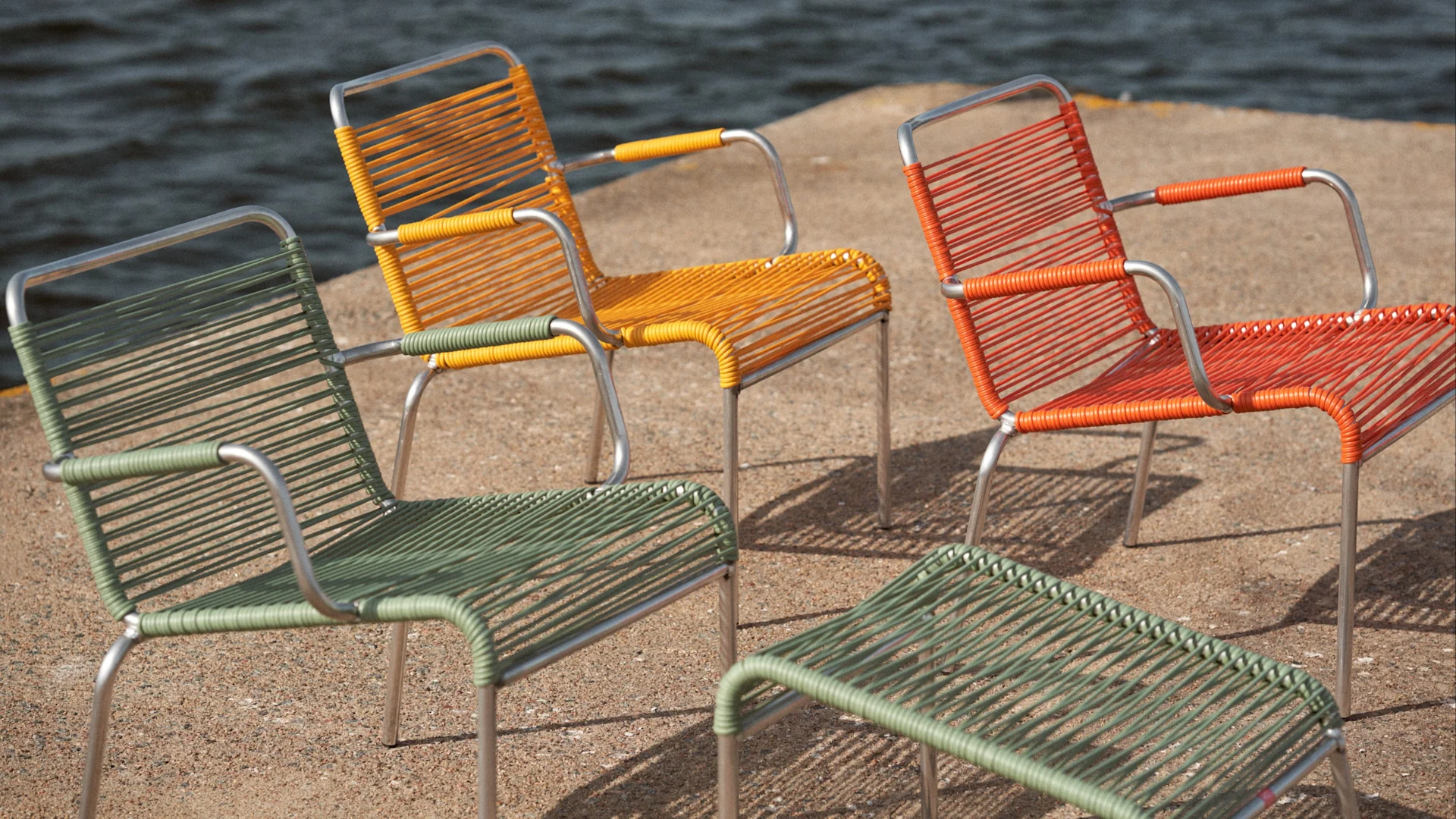 Tres sillas tejidas en verde, naranja y amarillo, con un taburete verde a juego, sobre arena junto al agua.
