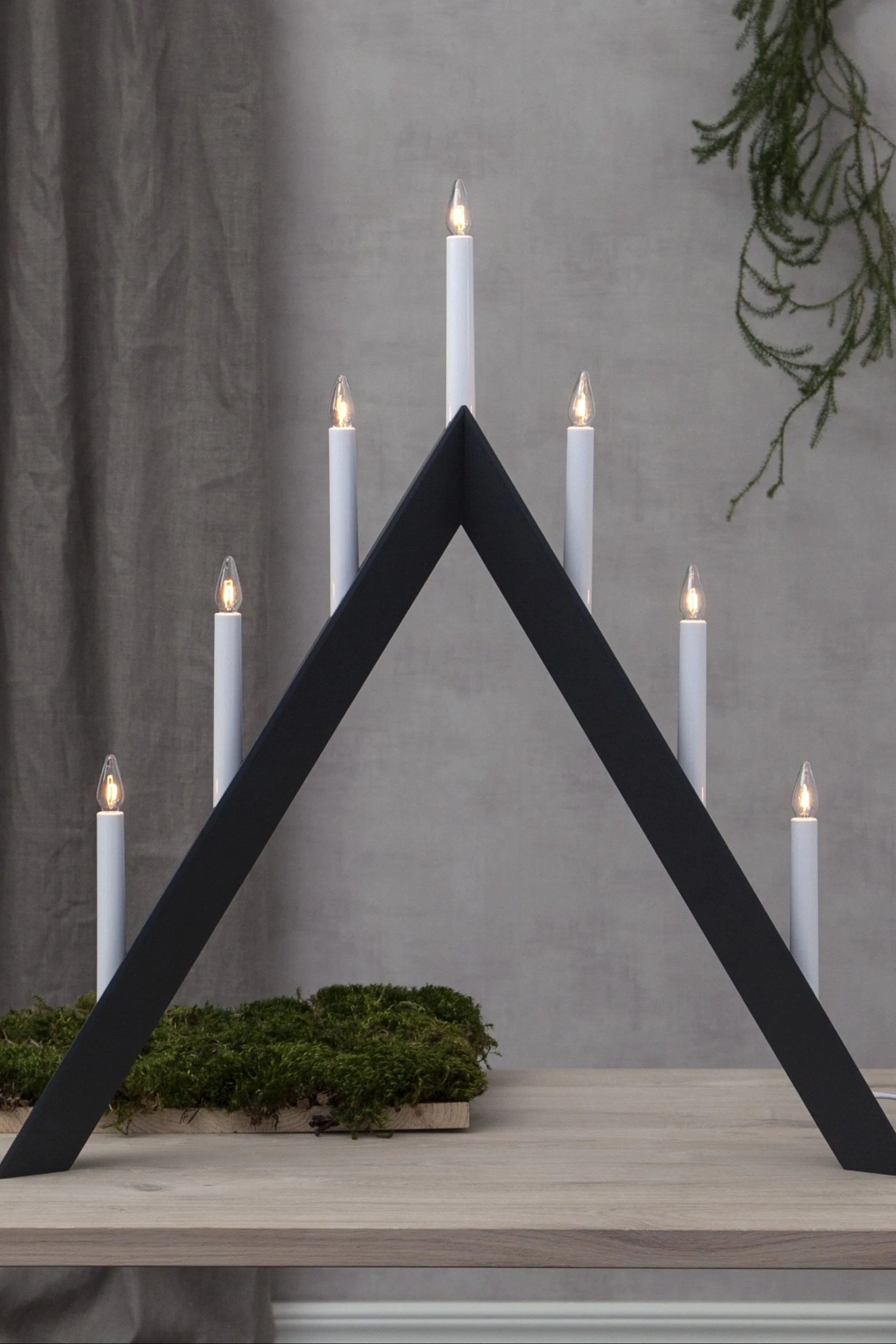 Un candelabro blanco y triangular con siete velas eléctricas encendidas sobre una superficie blanca, con un árbol de Navidad borroso al fondo.