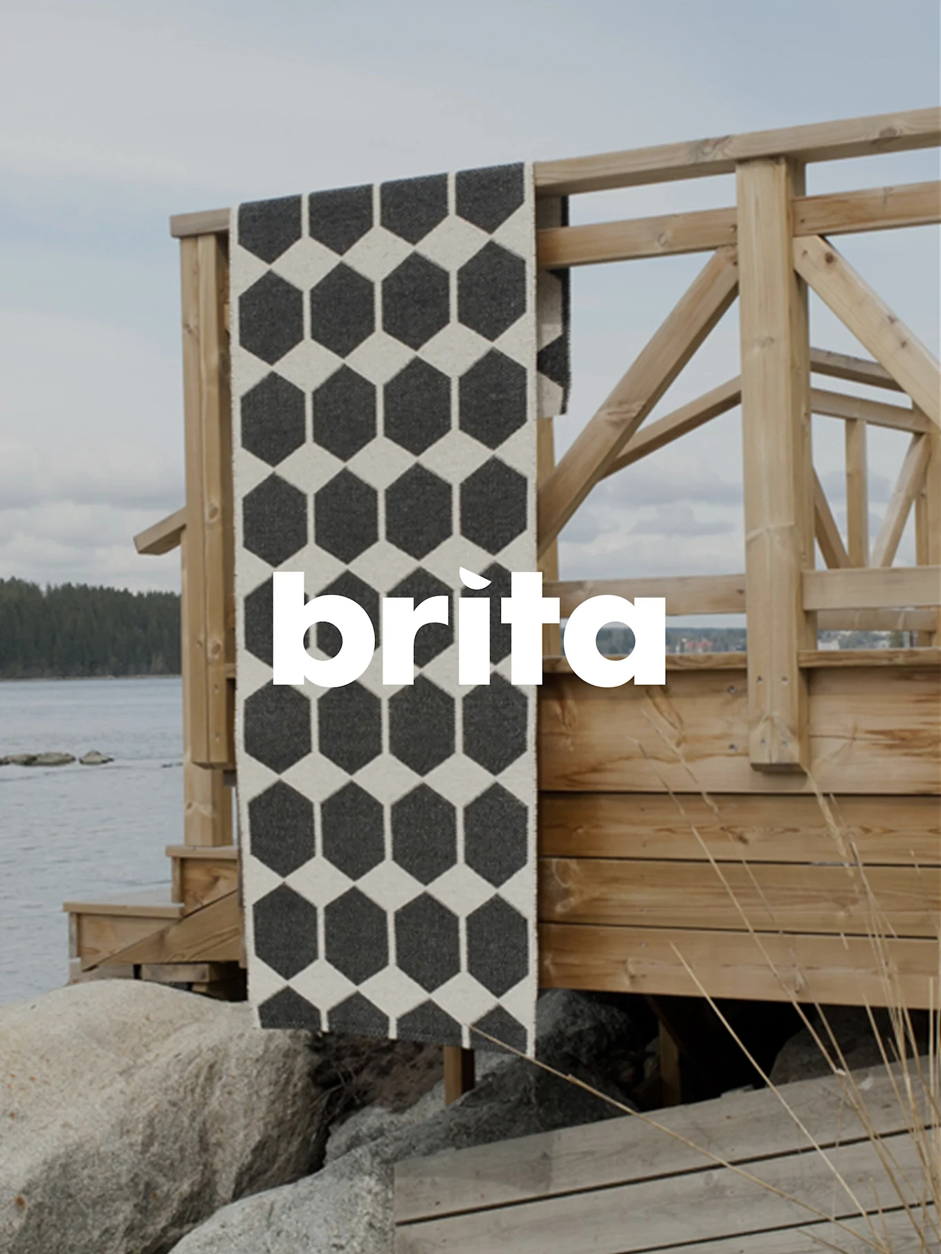 Una alfombra runner negra y blanquecina con patrón hexagonal, sobre una barandilla de madera junto al agua y rocas.