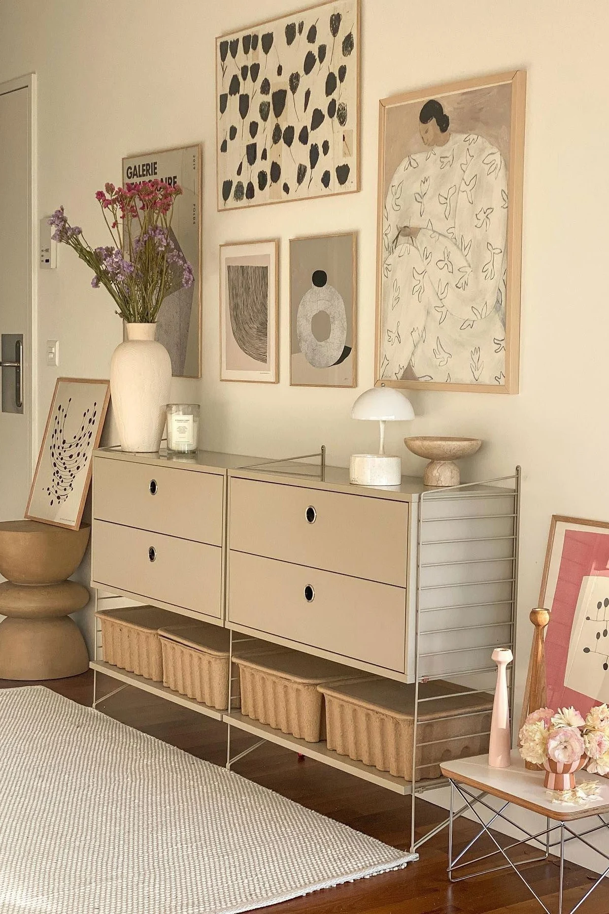 Una habitación moderna con un armario modular beige, cajones y cestas de almacenamiento. Un jarrón de flores, lámpara y arte abstracto adornan el espacio.