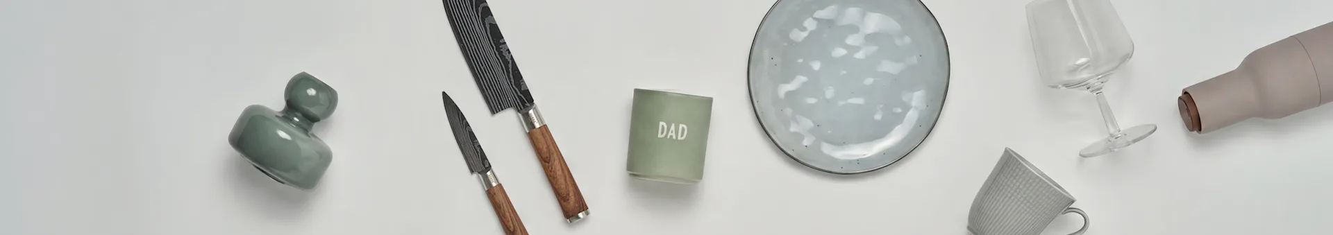 Una vista plana de artículos de cocina y regalo: un jarrón verde, dos cuchillos, una vela 'DAD', un plato de cerámica gris, una copa de vino, una botella beige y una taza gris.