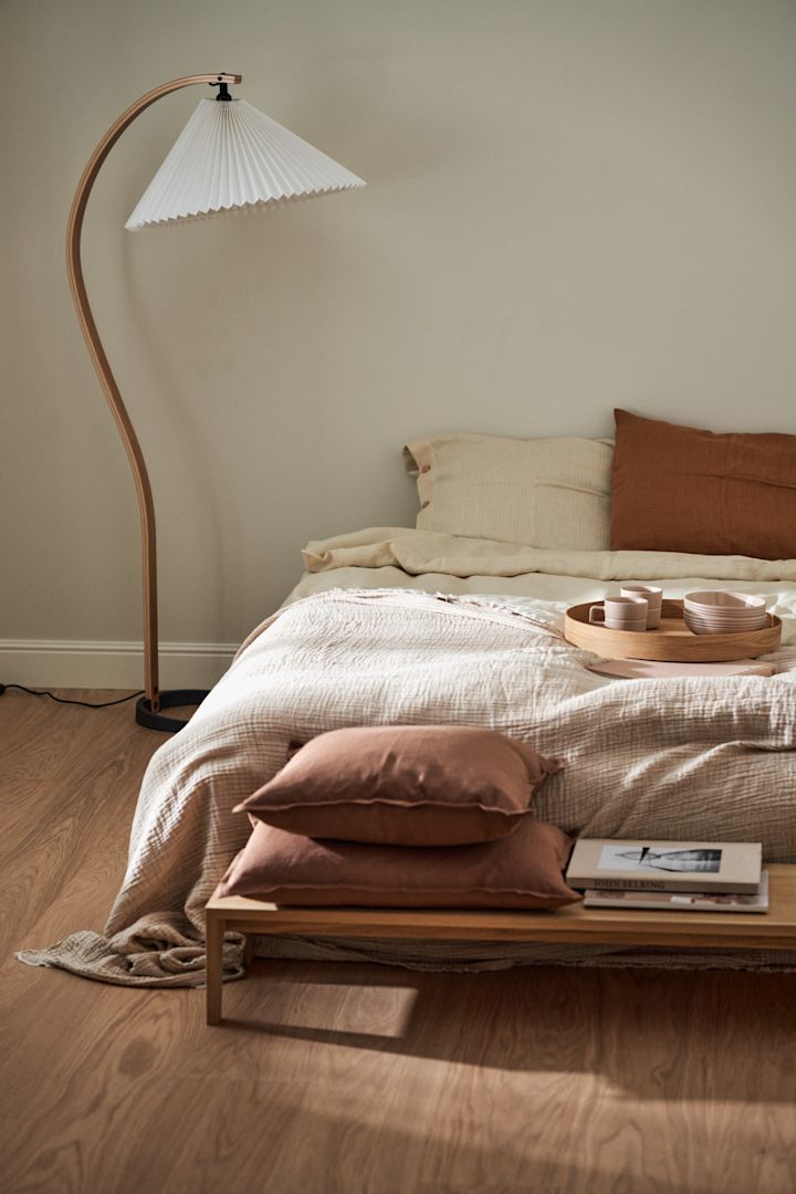 Aquí puedes ver un dormitorio en estilo Japandi con una cama, un banco frente a ella y la lámpara de pie Timberline de Gubi junto a ella.