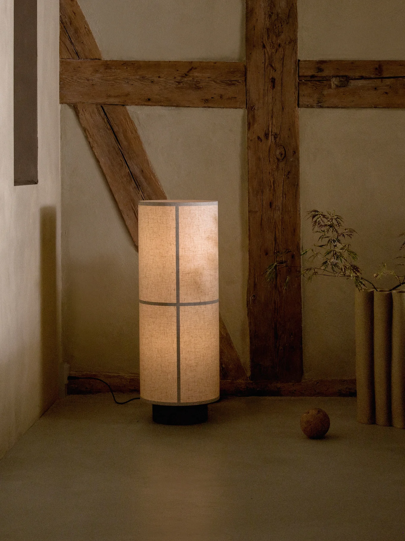 Lámpara de pie alta con pantalla de tela segmentada, proyectando un brillo cálido en una habitación rústica con vigas de madera expuestas.