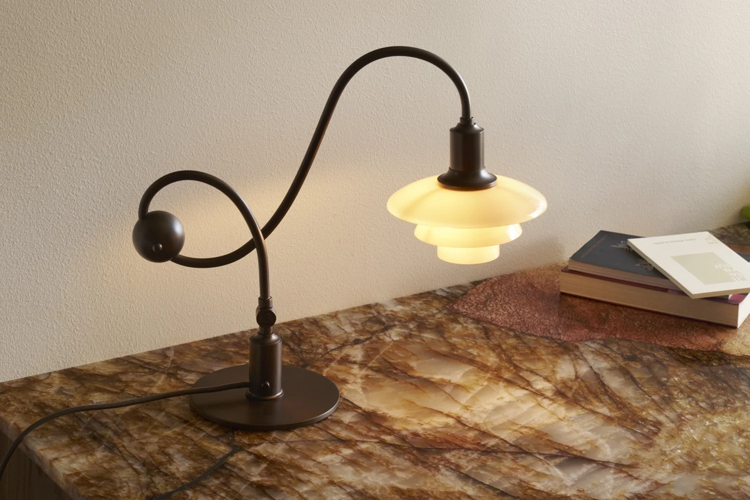 Lámpara de mesa de metal oscuro con tallo curvo y pantalla blanca escalonada y brillante sobre una superficie de mármol con libros.