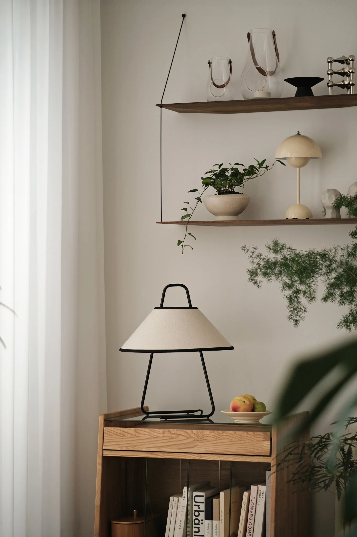 Sala de estar moderna con lámpara de mesa crema y negra sobre un mueble de madera clara, estantes decorativos y plantas.
