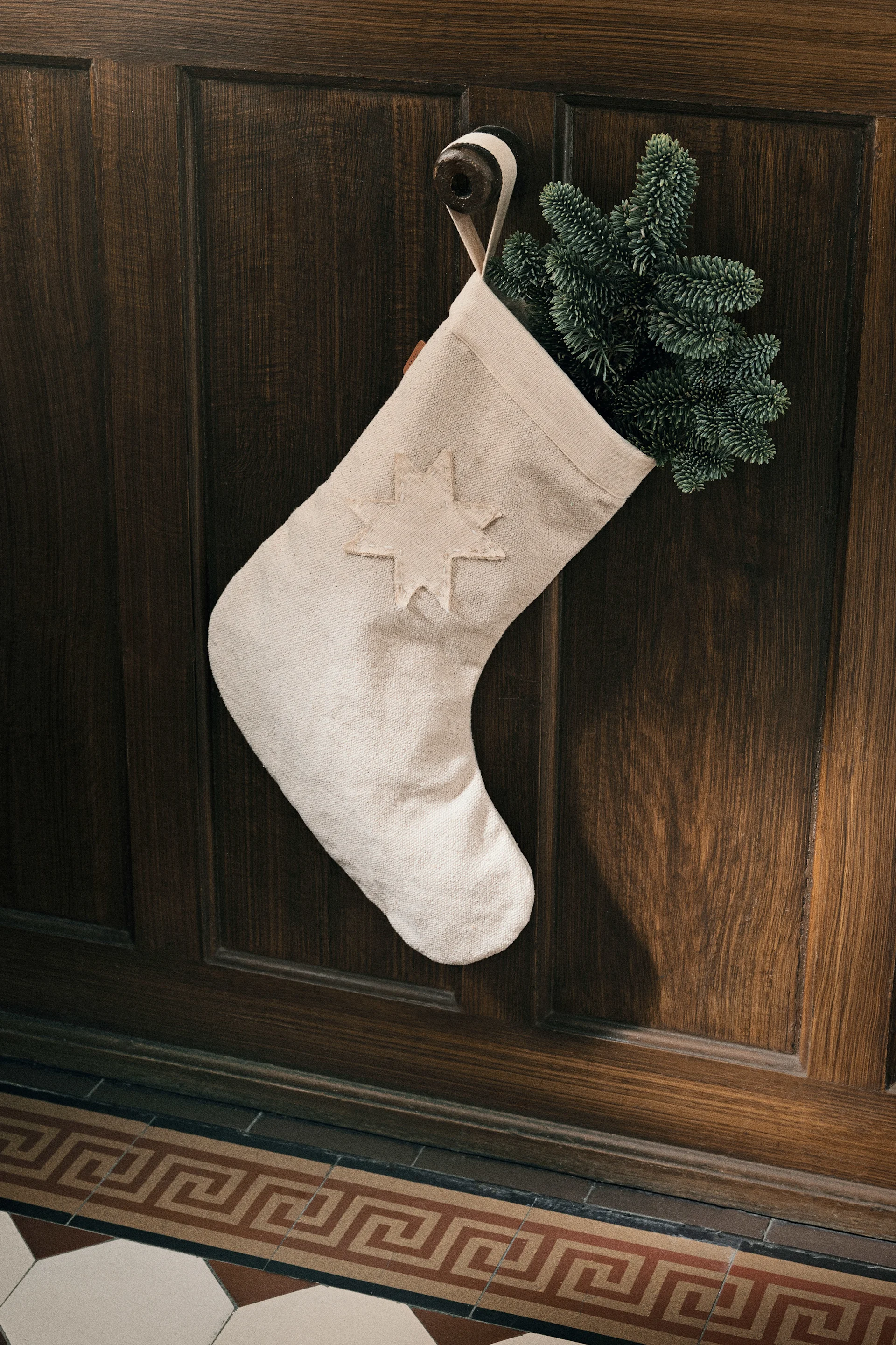 Manualidades navideñas con ramas de abeto: aquí se ve el calcetín de Navidad Vela de ferm LIVING, adornado con ramas de abeto, colgado de un gancho en la pared.