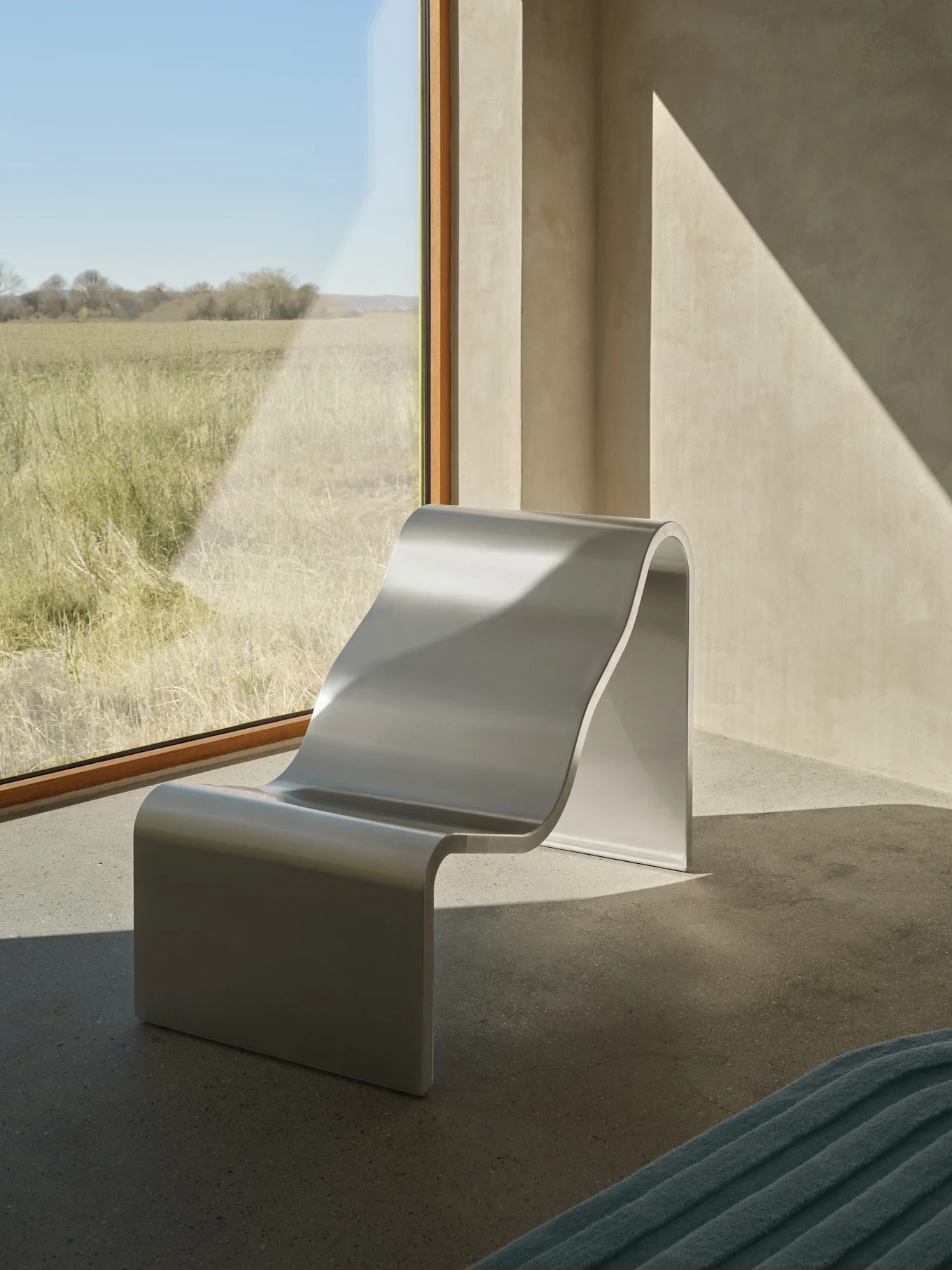 Una moderna silla de salón plateada en forma de S se encuentra junto a un gran ventanal con vistas a un campo soleado, con patrones diagonales de luz solar en las paredes.