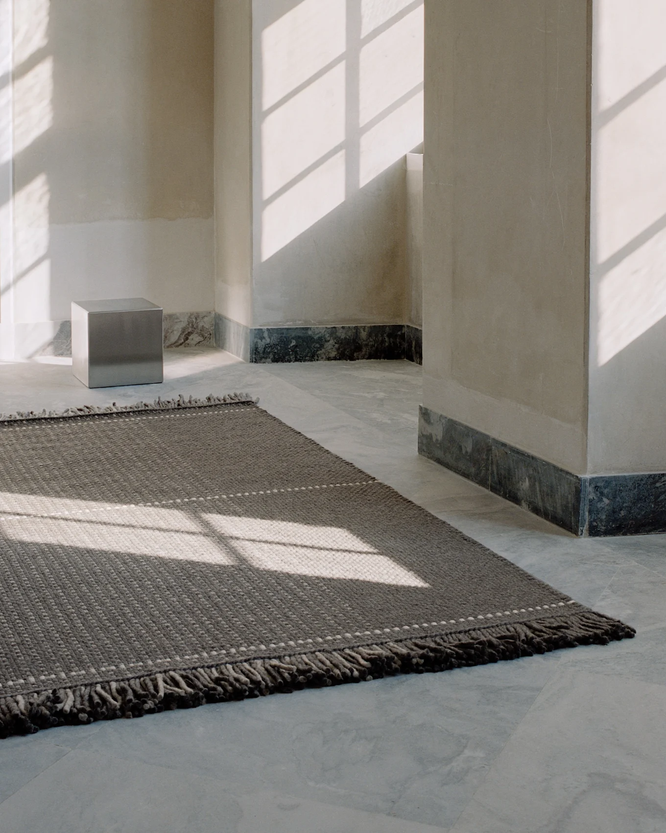 Una alfombra gris oscuro texturizada con puntos claros en un suelo de baldosas claras, iluminada por sombras de ventana en una habitación minimalista.